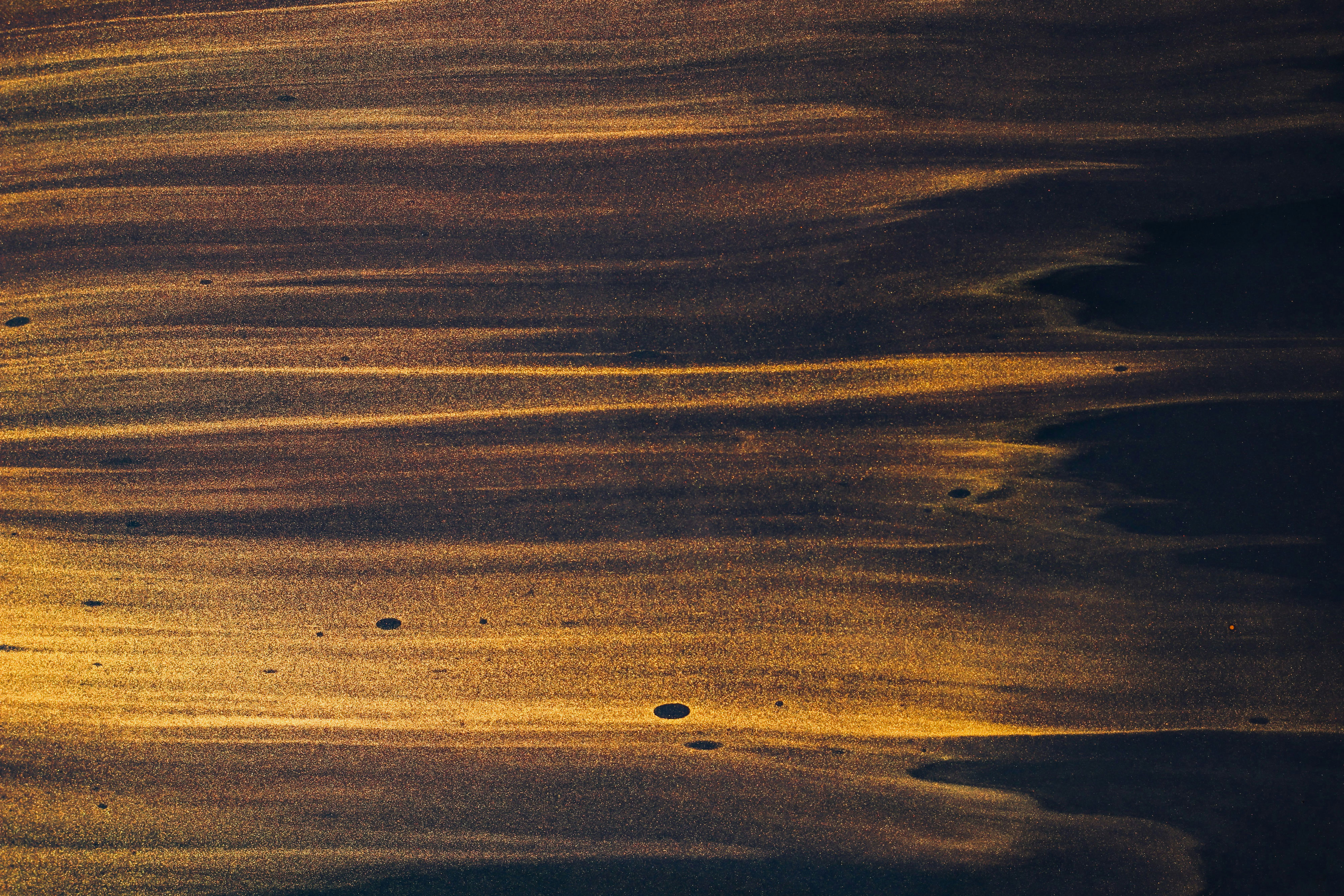 Abstraktes Bild von dunklem Sand mit goldenen Lichtstreifen, die ein wellenförmiges, strukturiertes Muster bilden. Auf der sandigen Oberfläche sind vereinzelte dunkle Flecken zu erkennen.
