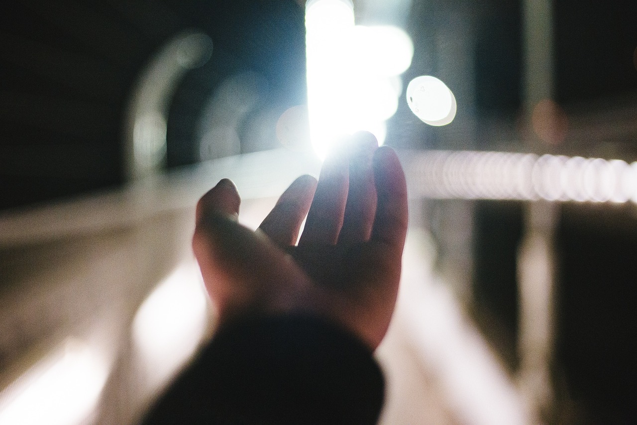 Eine Hand streckt sich nachts auf ein helles, verschwommenes Licht am Ende eines Tunnels oder Weges aus, vor dem Hintergrund unscharfer Geländer und einer dunklen Umgebung.