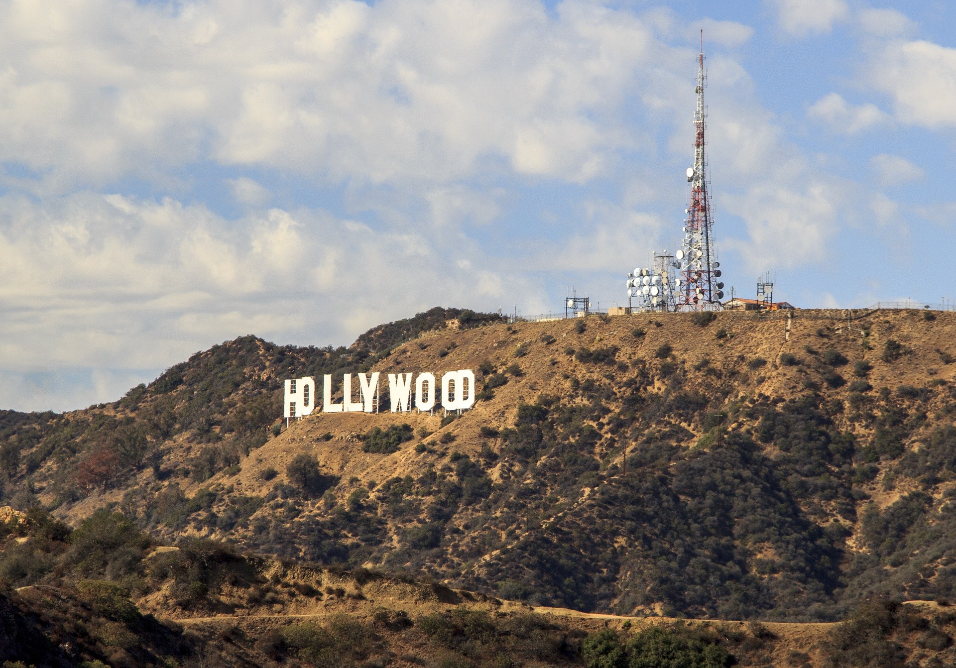 Das Hollywood-Zeichen auf einem braunen Hügel in Los Angeles, in dessen Nähe sich ein großer Funkturm und Antennen befinden. Darüber erstreckt sich ein blauer Himmel mit vereinzelten Wolken.