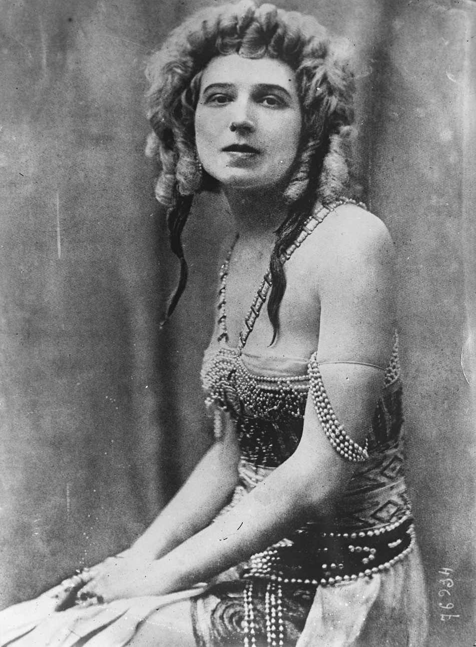 The picture shows a historical black and white photograph of Ida Rubinstein in an elaborately decorated stage costume with pearls. She sits with folded hands and voluminous curly hair in front of a neutral background.