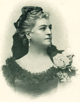 Schwarz-Weiß-Porträt von Lilli Lehmann in förmlicher Kleidung mit lockigem Haar, die Ohrringe und eine große Blumenverzierung auf ihrem Kleid trägt und leicht zur Seite schaut.
