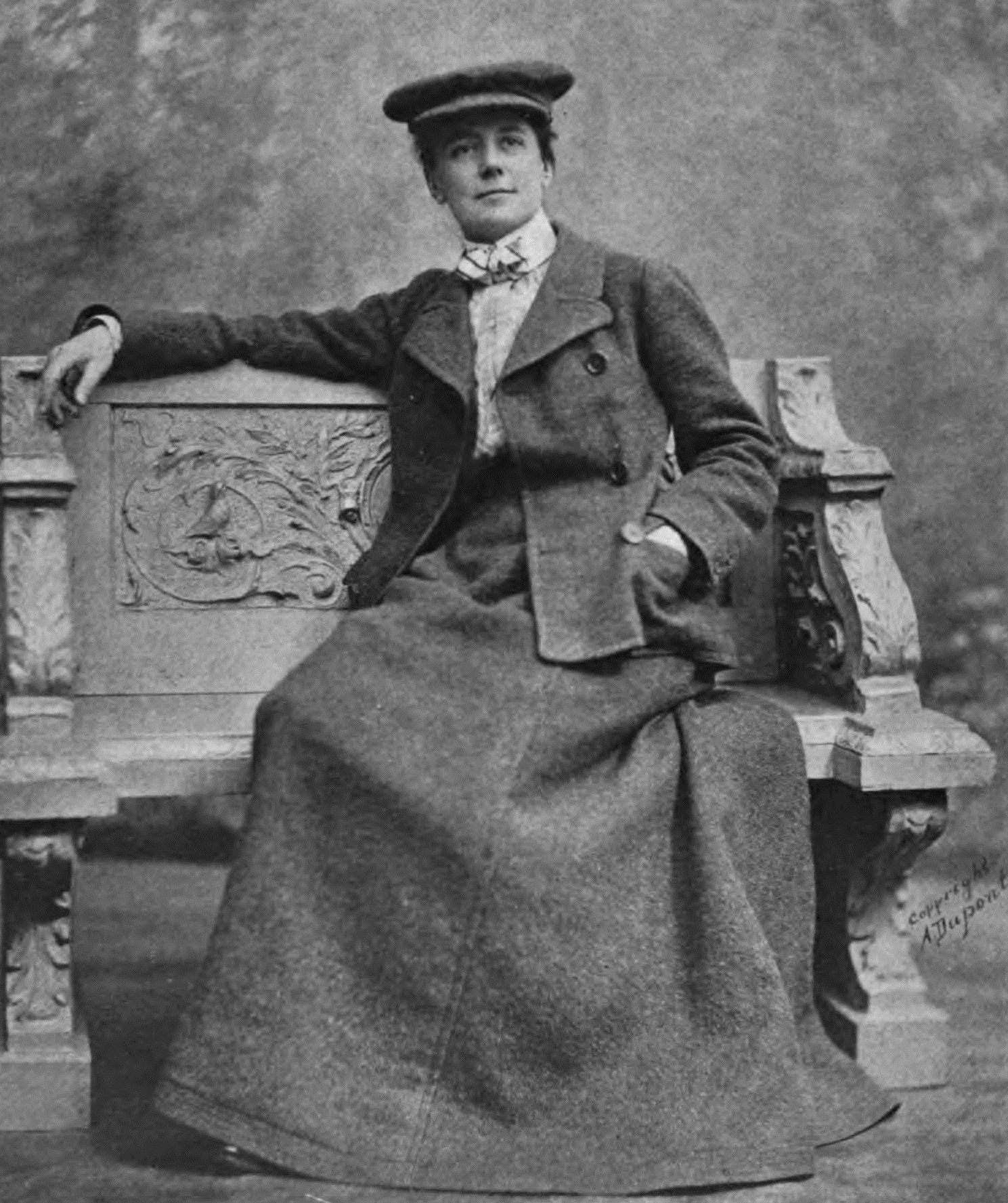 Ethel Smyth sits casually on an ornate wooden bench in a large woollen skirt and jacket. She is wearing a cap and looks confident.
