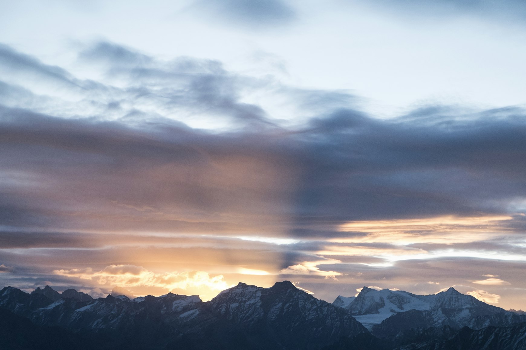 Verschneite Berggipfel unter einem dramatischen Himmel, an dem bei Sonnenaufgang oder Sonnenuntergang Sonnenstrahlen und Schatten durch bunte Wolken brechen.