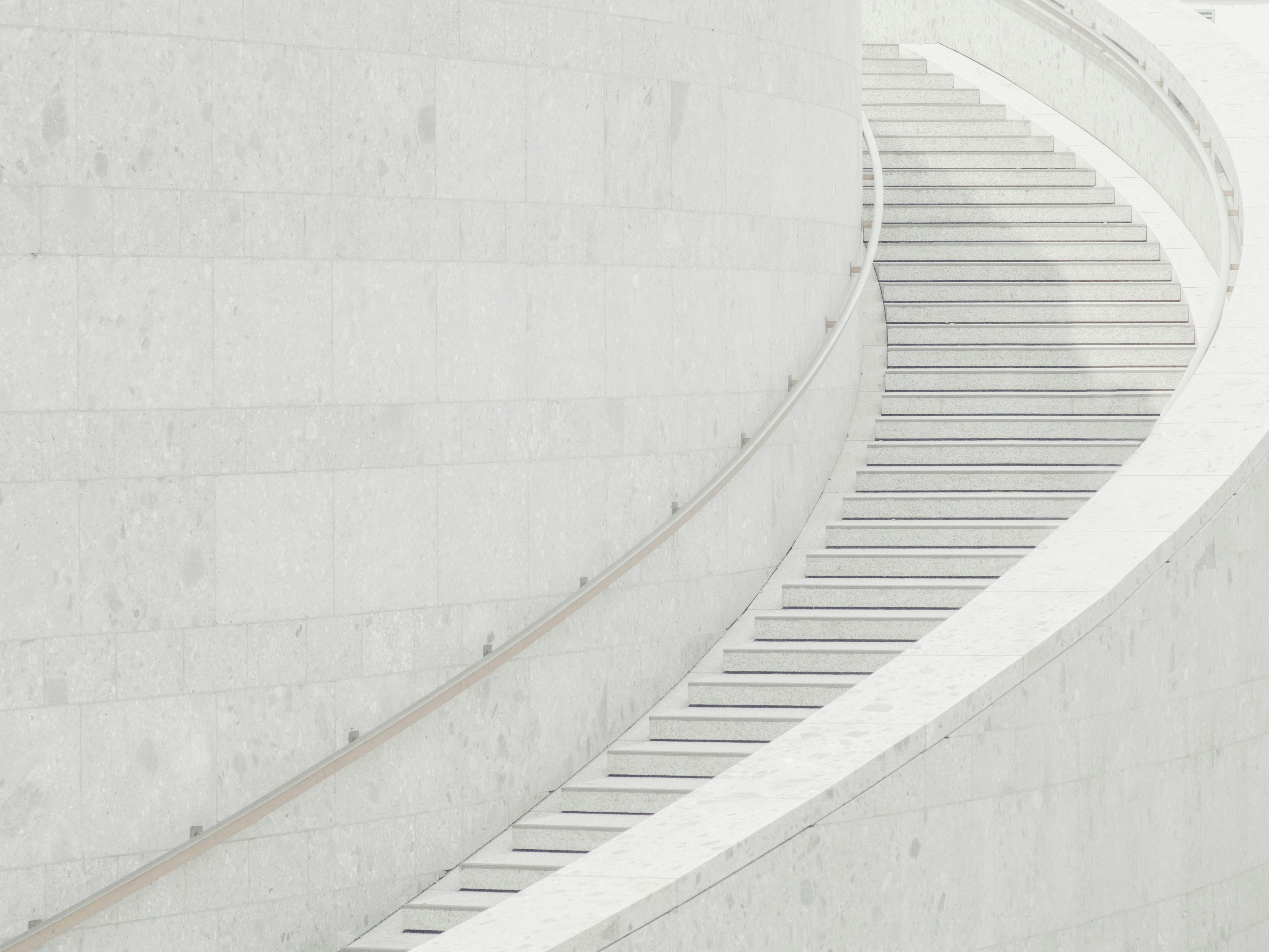 Eine geschwungene Treppe mit weißen Steinstufen und einem glatten Geländer schlängelt sich entlang einer großen, hellen Steinmauer in einem modernen, minimalistischen architektonischen Umfeld.