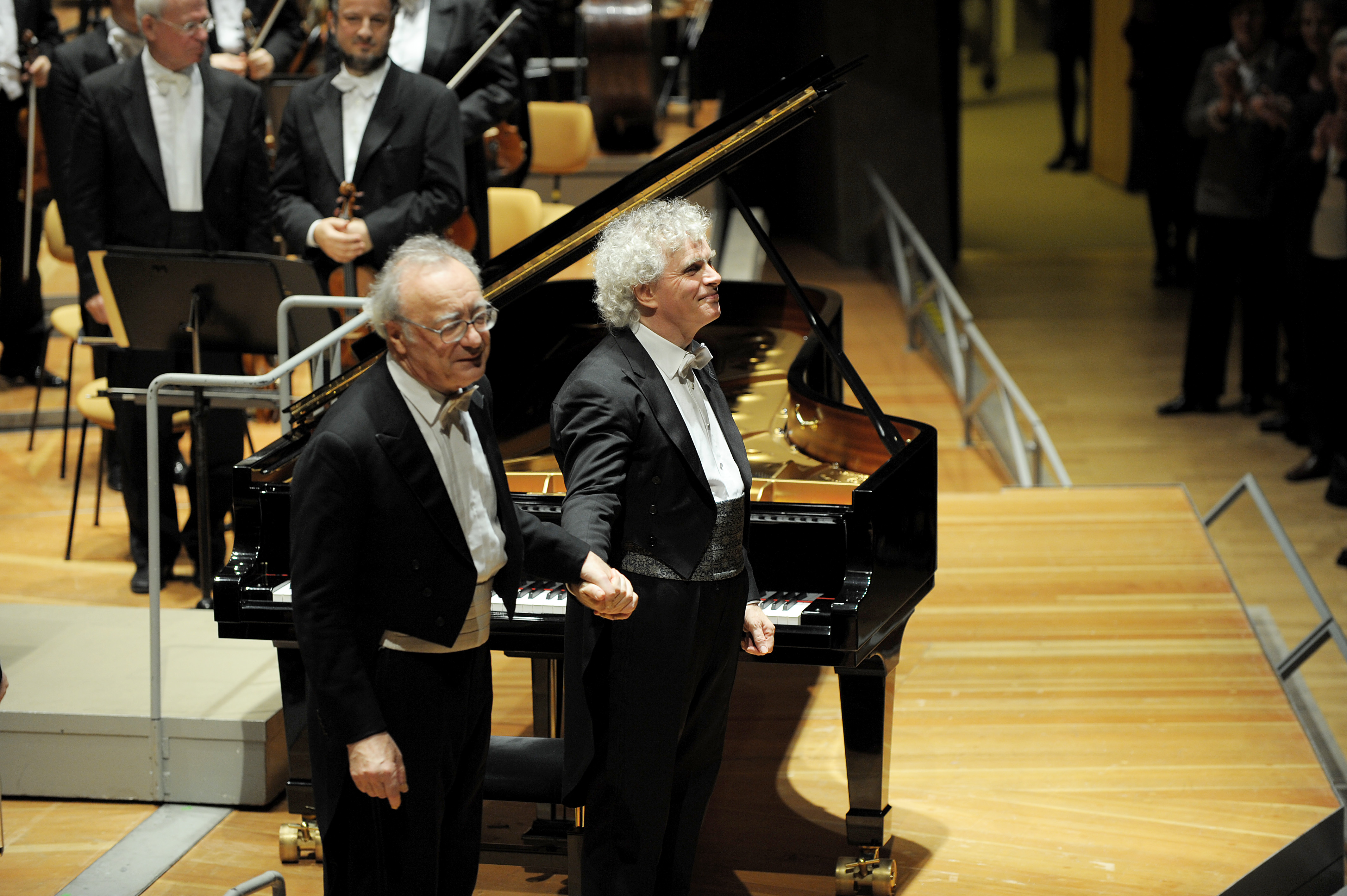 After a performance, Alfred Brendel and Simon Rattle stand holding hands in front of a grand piano on the concert stage of the Berlin Philharmonic Hall, with an orchestra and audience visible in the background.