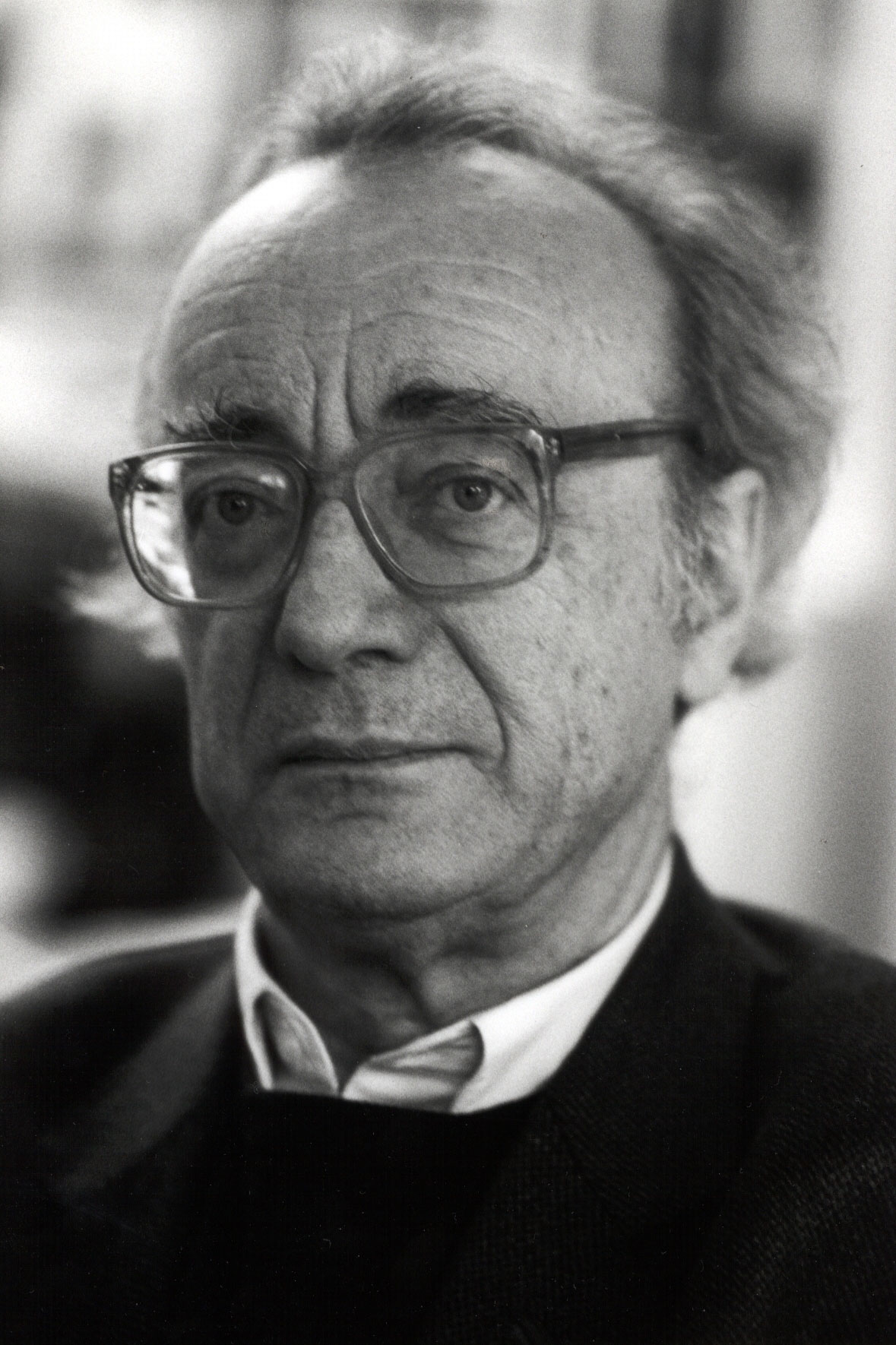 Black-and-white portrait of Alfred Brendel with thinning hair, large glasses and a serious expression. He is wearing a suit jacket, jumper and collared shirt. The background is slightly out of focus.