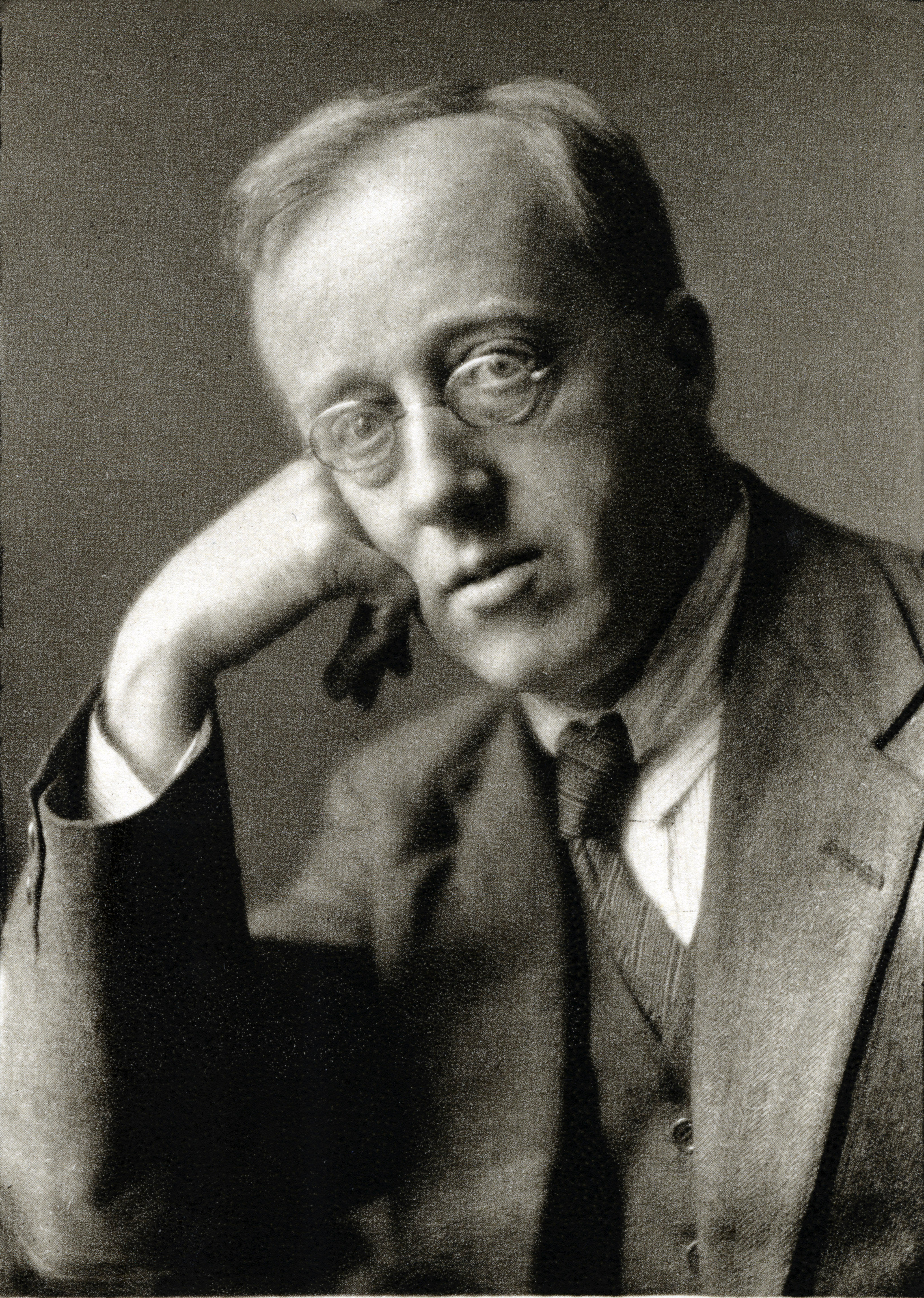 Schwarz-weiß-Fotografie von Gustav Holst, er trägt Anzug, Kravatte und eine Brille und lehnt seinen Kopf auf eine Hand.