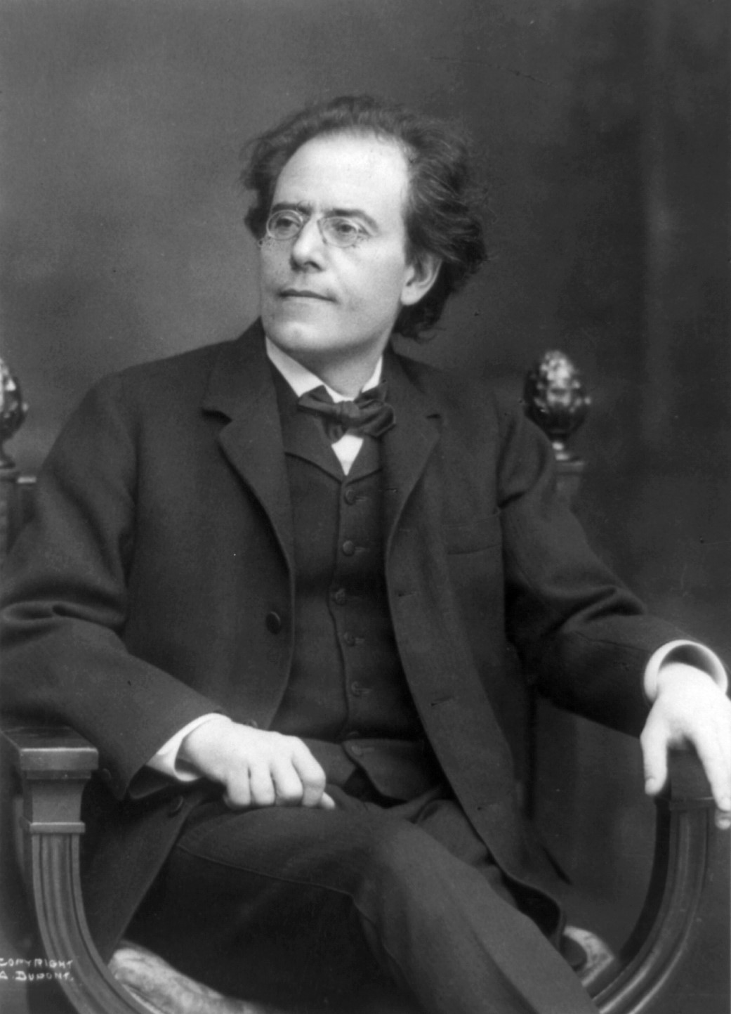 Gustav Mahler mit welligem Haar, Brille und Anzug sitzt in einem verzierten Stuhl und blickt mit ruhigem Gesichtsausdruck leicht zur Seite. Das Bild ist schwarz-weiß vor einem neutralen Hintergrund.
