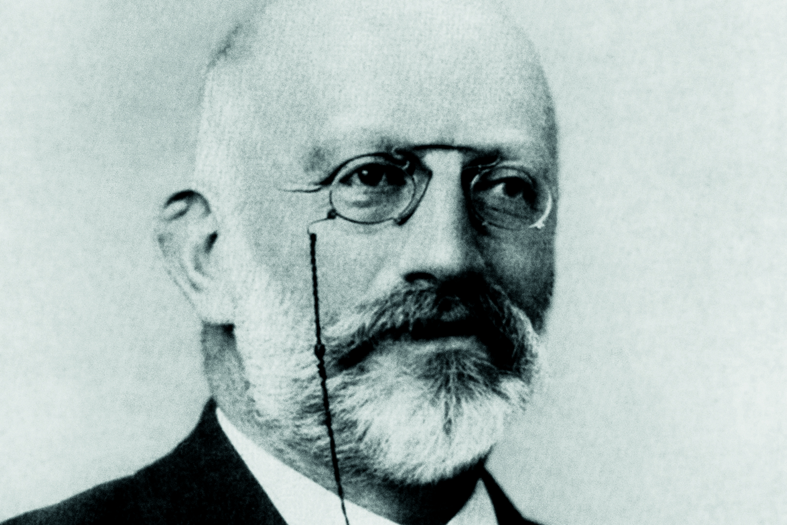 Schwarz-Weiß-Porträt von Hermann Wolff mit Glatze, runder Brille, Schnurrbart und Vollbart. Er trägt einen Anzug und hat ein Monokel, das mit einer dünnen Schnur an seiner Brille befestigt ist.