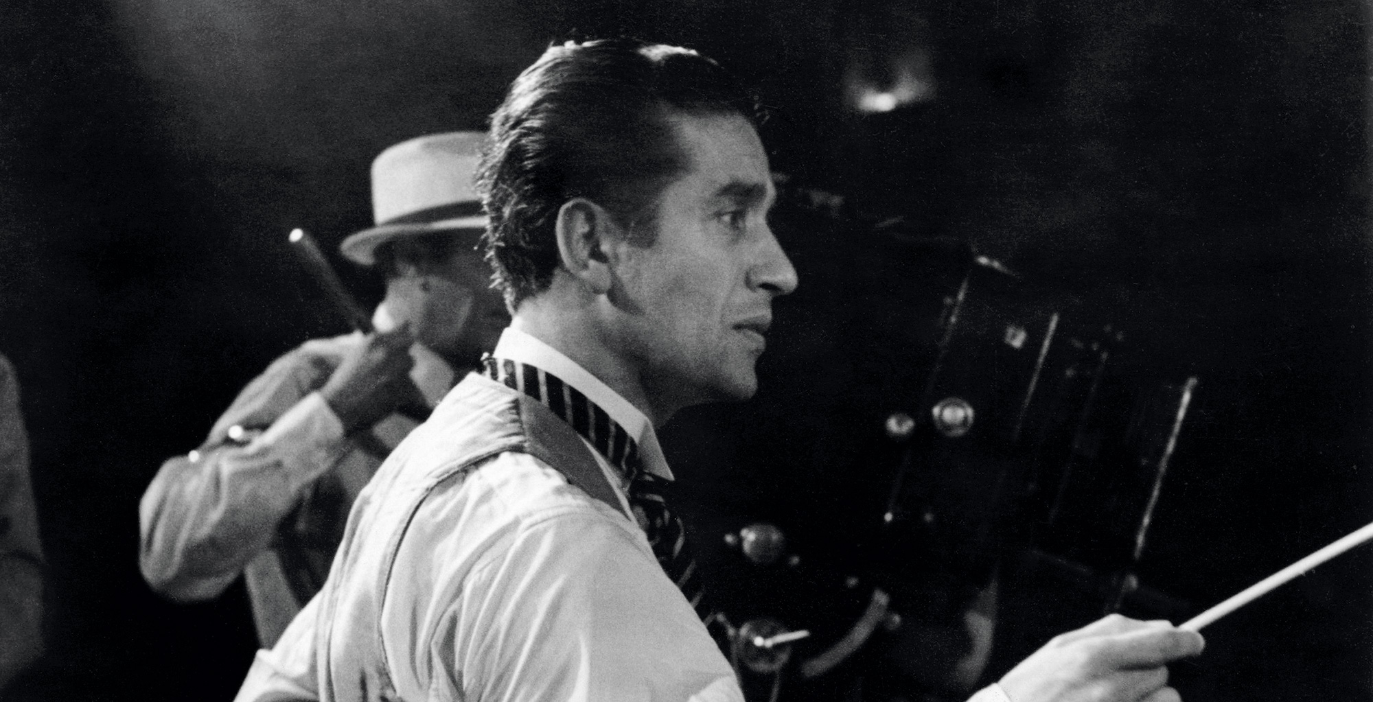 Sergiu Celibidache in a dress shirt and tie holds a conductors baton, concentrating intently. A person in a hat and a film camera are visible in the dimly lit background.
