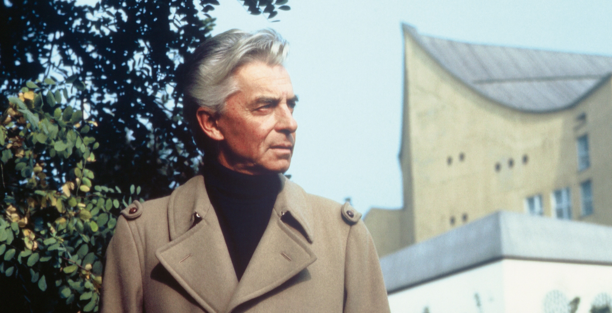 Herbert von Karajan mit grauem Haar, der einen hellbraunen Trenchcoat und einen schwarzen Rollkragenpullover trägt, steht im Freien in der Nähe von belaubten Ästen, wobei im Hintergrund ein modernes Gebäude mit einem geschwungenen Dach zu sehen ist.