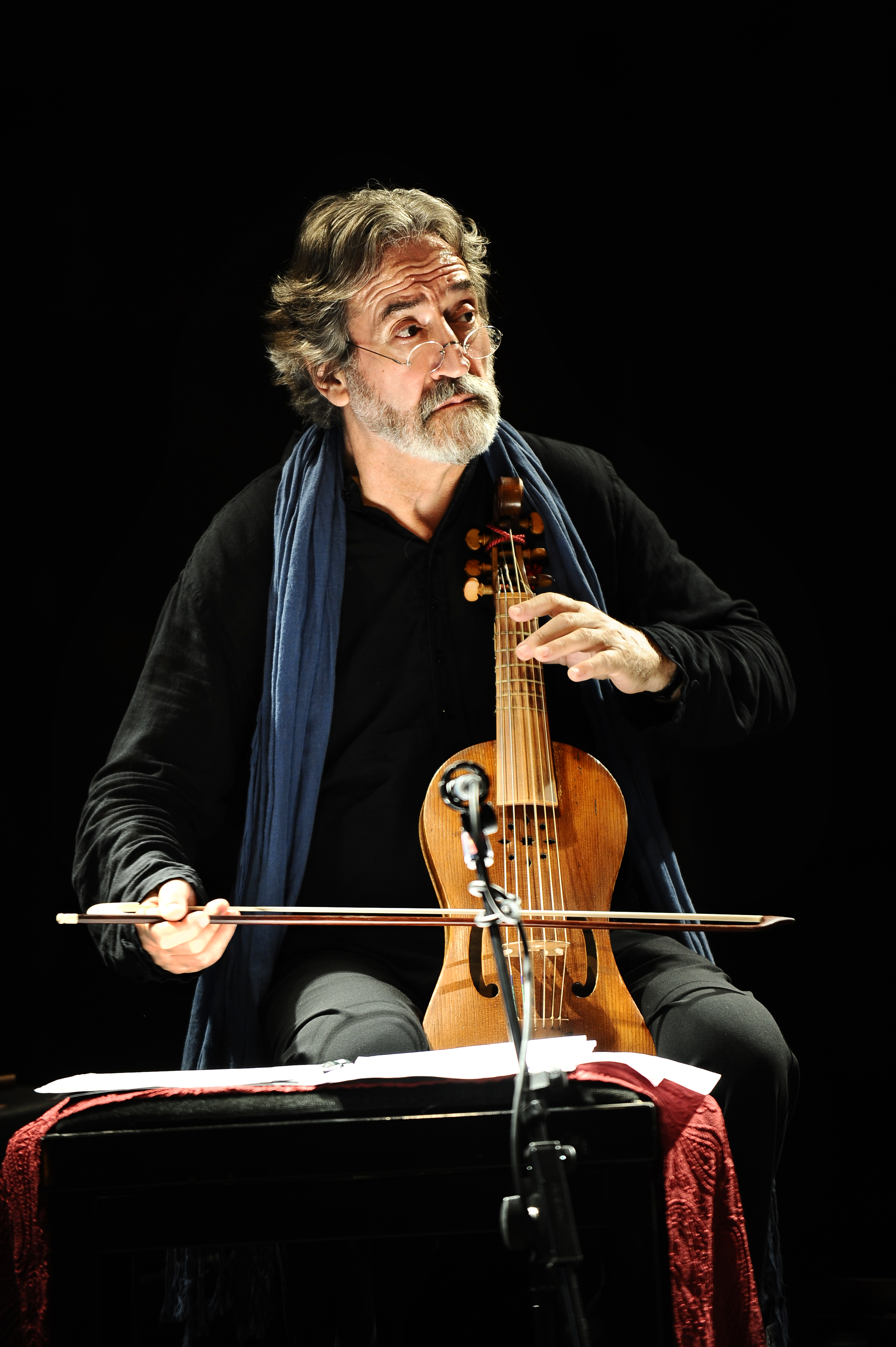 A bearded man with gray hair, wearing a dark shirt and blue scarf, plays a bowed string instrument on stage, focused intently, with sheet music in front of him and a microphone nearby.