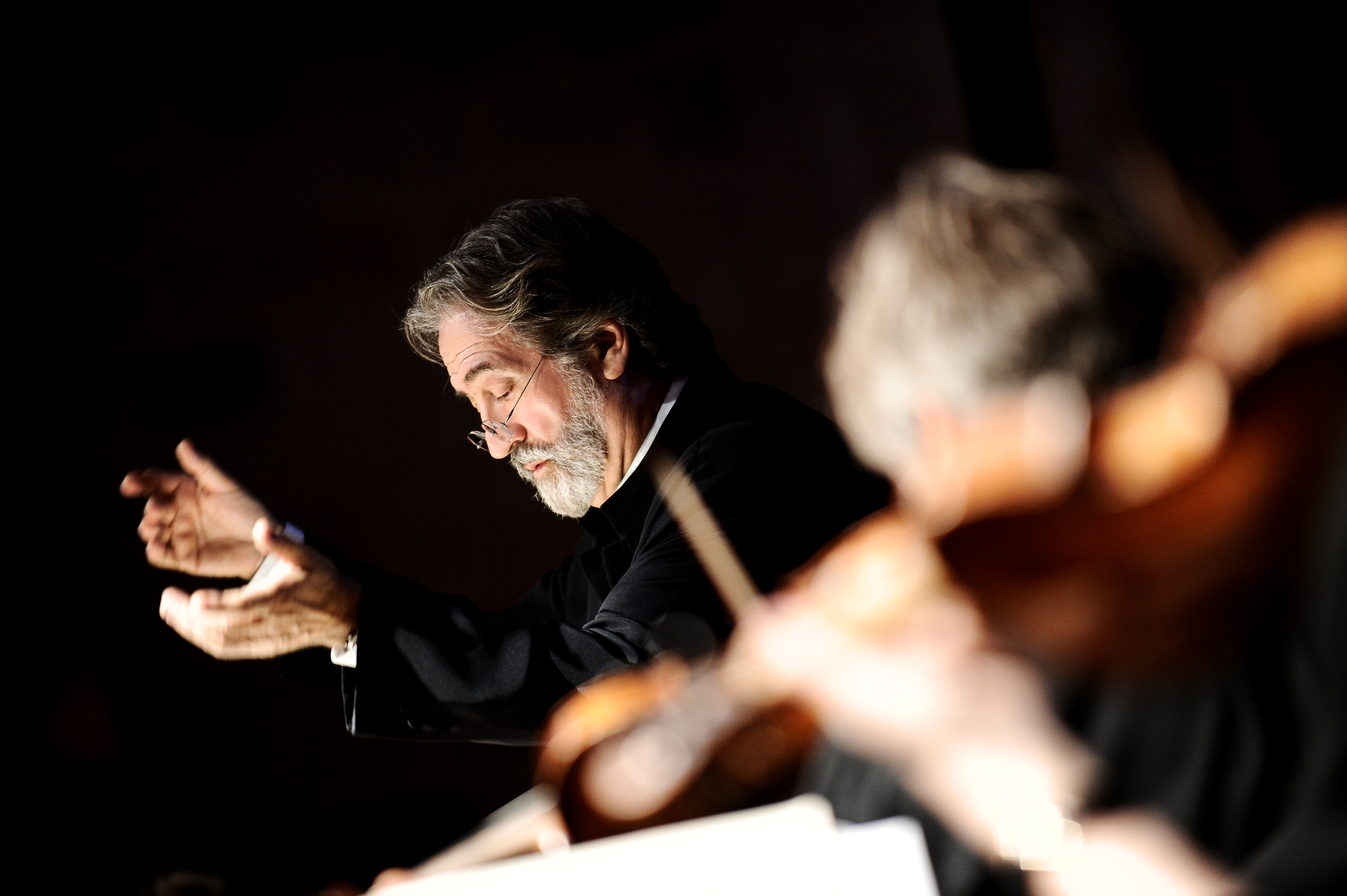 A bearded man in glasses conducts an orchestra, captured mid-motion with expressive hands. The foreground is blurred, showing musicians and string instruments out of focus.