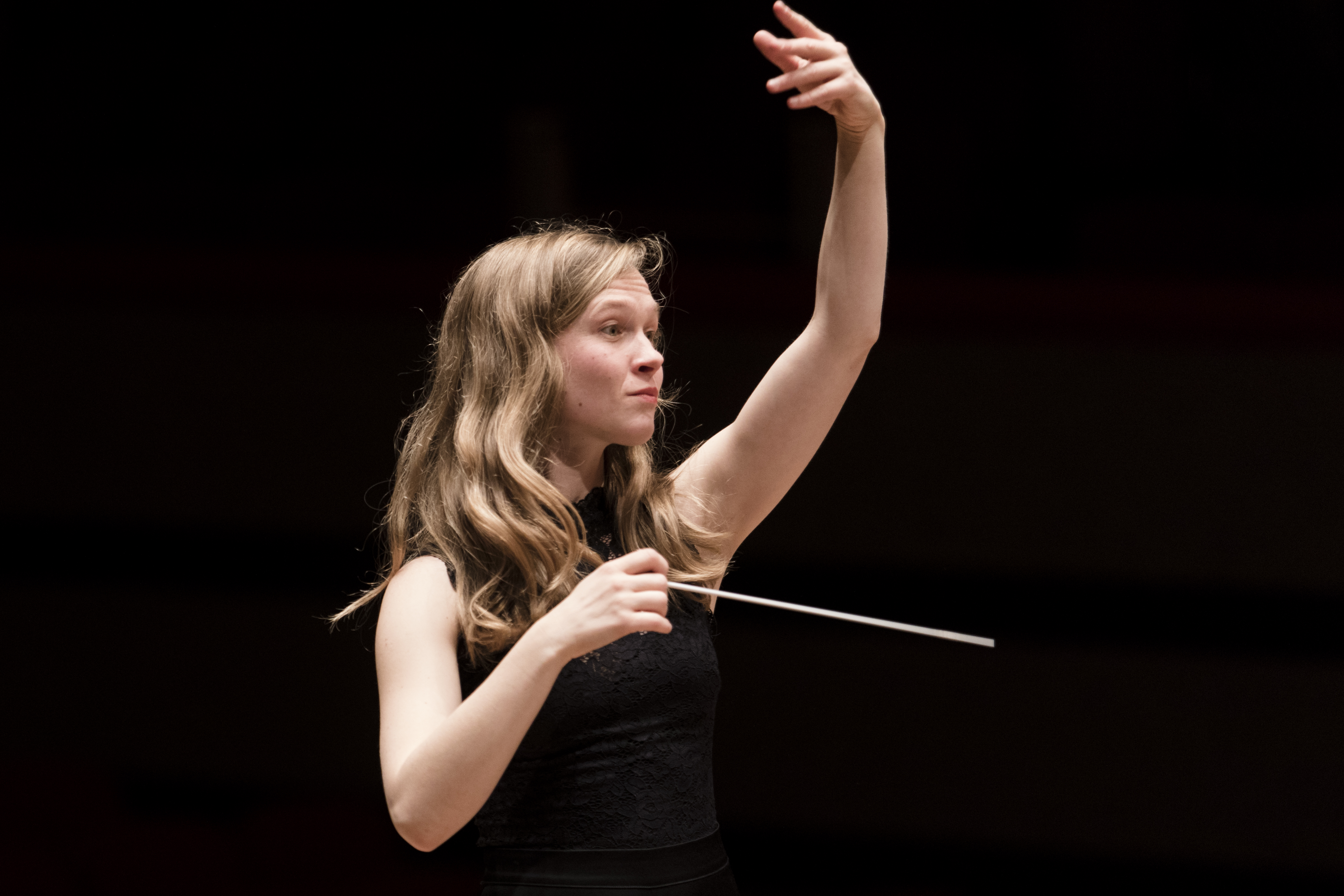 Eine Frau mit langen Haaren dirigiert ein Orchester, hält einen Taktstock in einer Hand und hebt die andere Hand. Sie trägt ein ärmelloses schwarzes Oberteil vor einem dunklen Hintergrund.