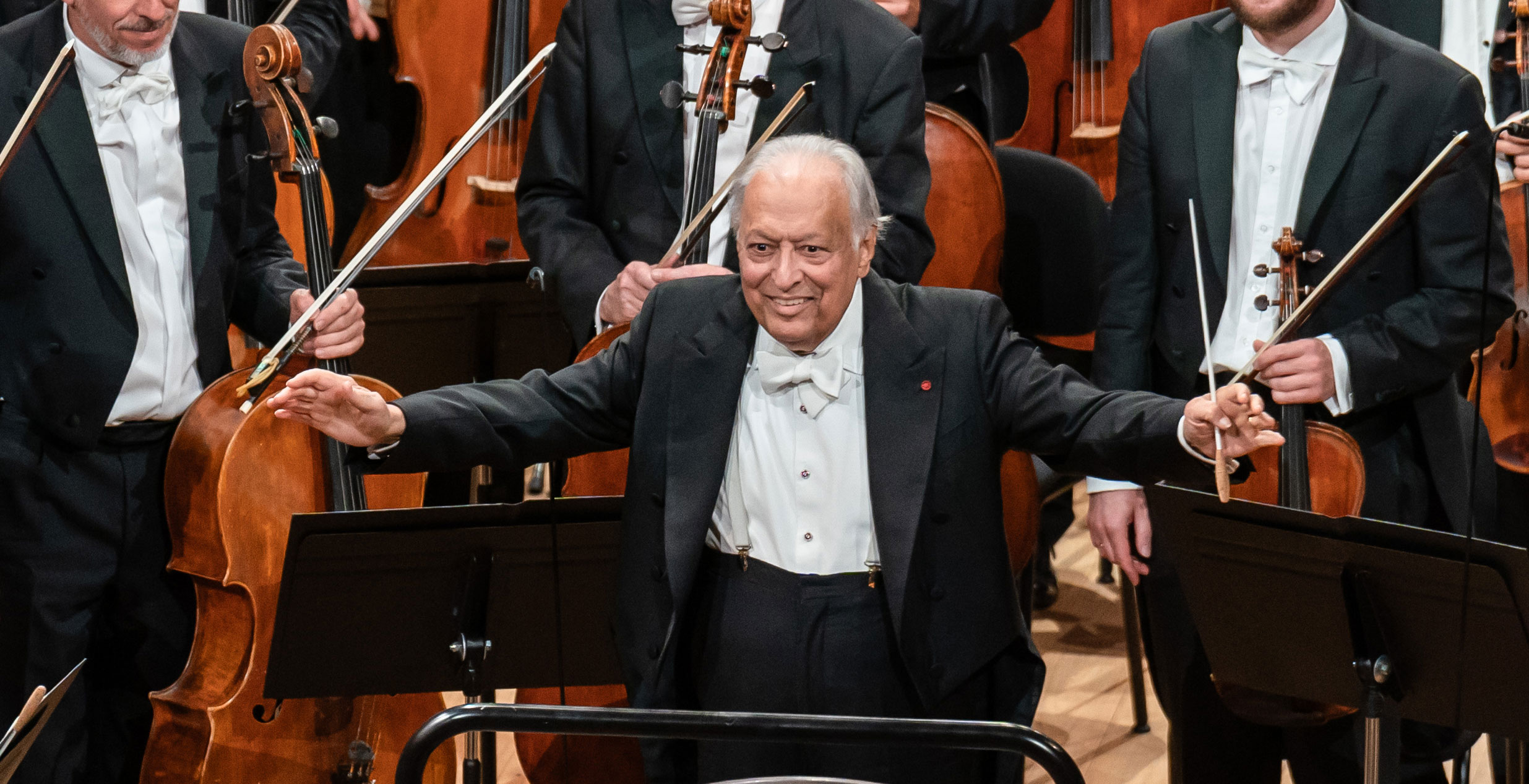 Ein älterer männlicher Dirigent in förmlicher Kleidung steht lächelnd mit erhobenen Armen, einen Taktstock haltend, vor einem Orchester mit Musikern und Streichinstrumenten hinter ihm.
