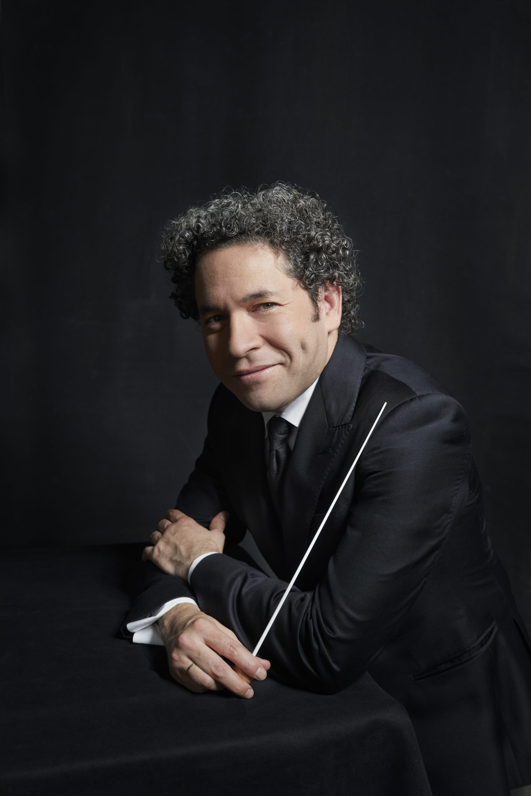 Gustavo Dudamel with curly hair in a suit leans against a dark table and holds a baton in front of a dark background. He has dimples and looks directly into the camera.