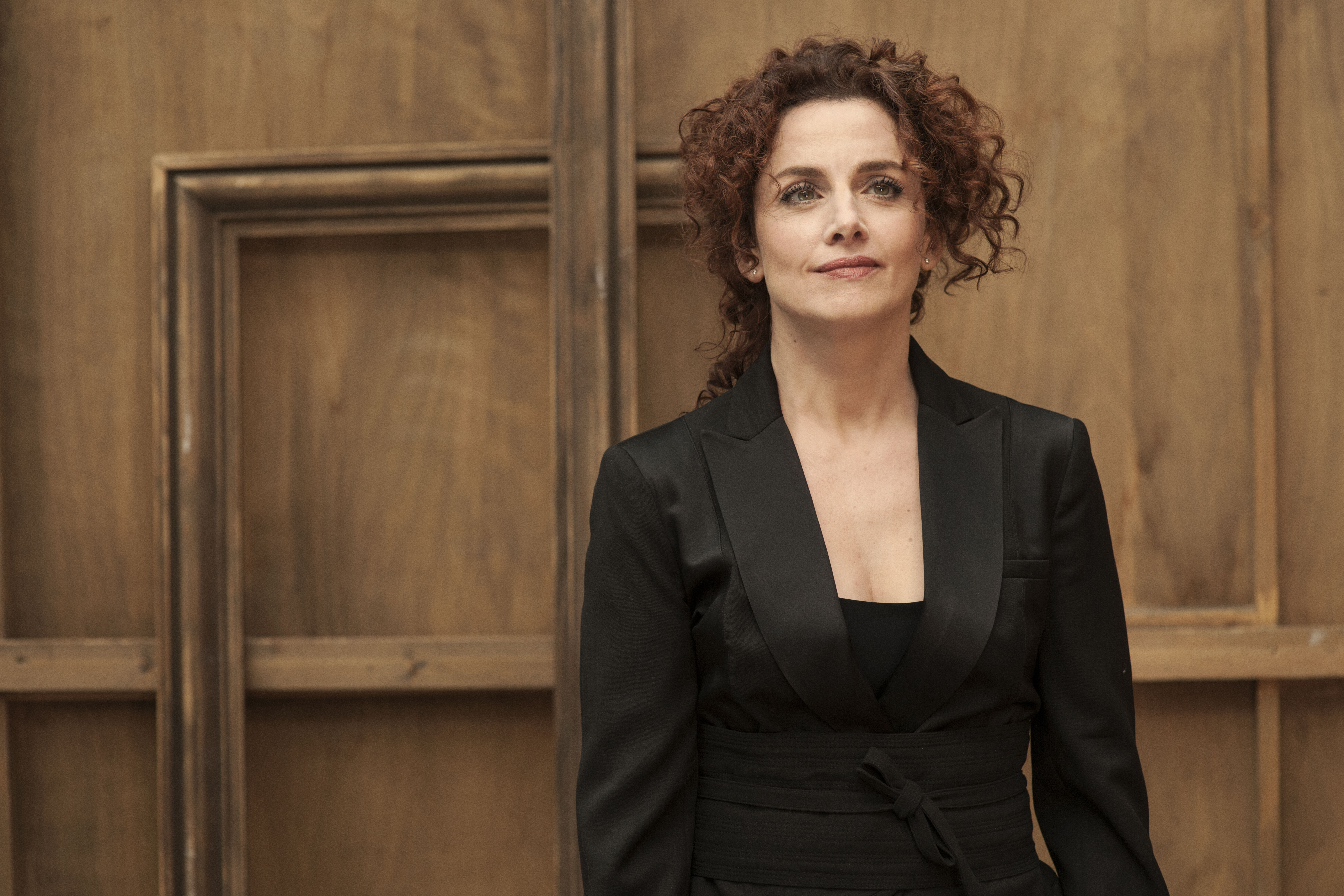 A woman with reddish curls looks calmly into the camera, wearing a long black jacket and standing in front of a wood-panelled wall.