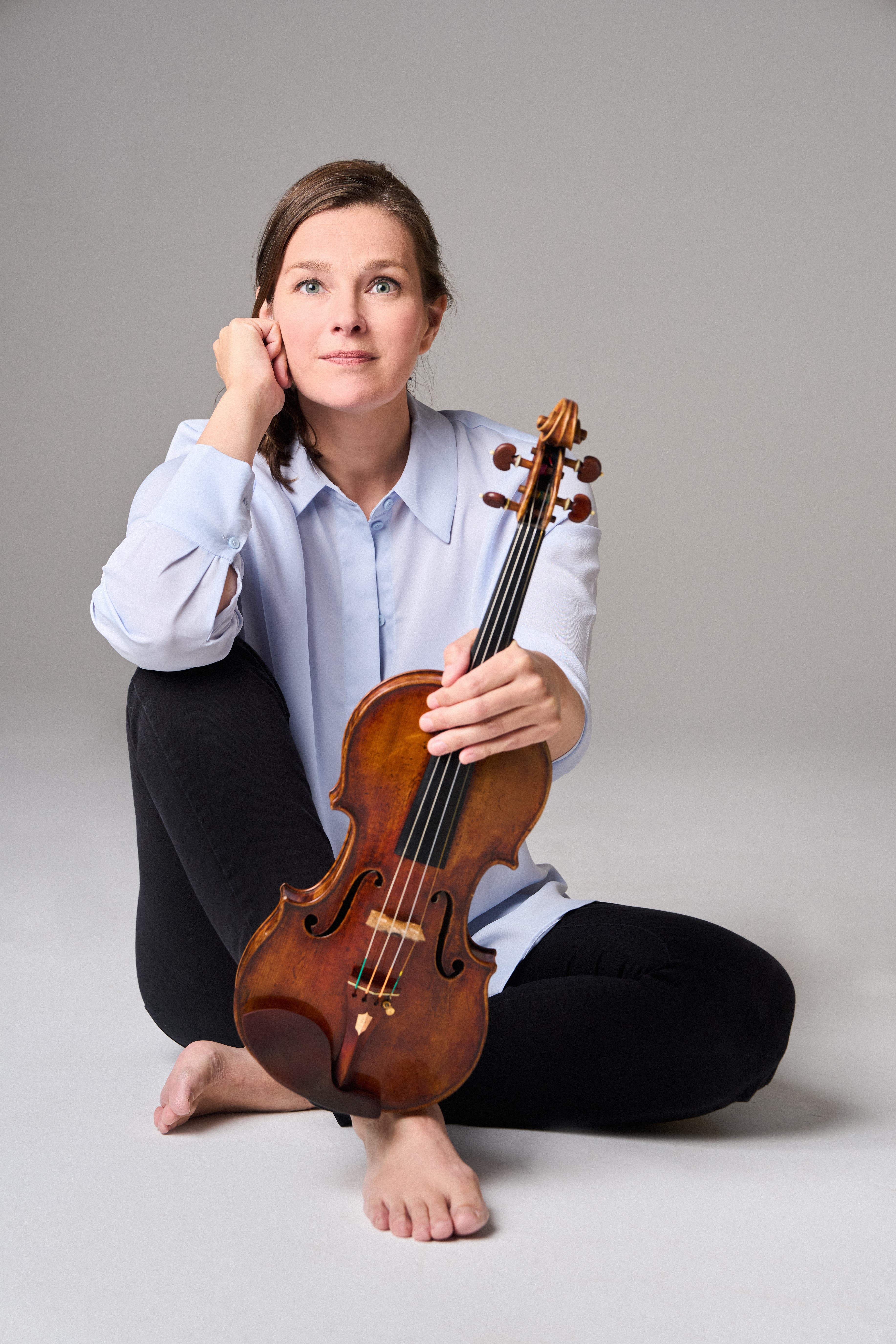 Janine Jansen mit langen Haaren, einem hellblauen Hemd und einer schwarzen Hose sitzt barfuß auf dem Boden. Sie hält eine Geige senkrecht auf ihrem Fuß und blickt nachdenklich in die Kamera.
