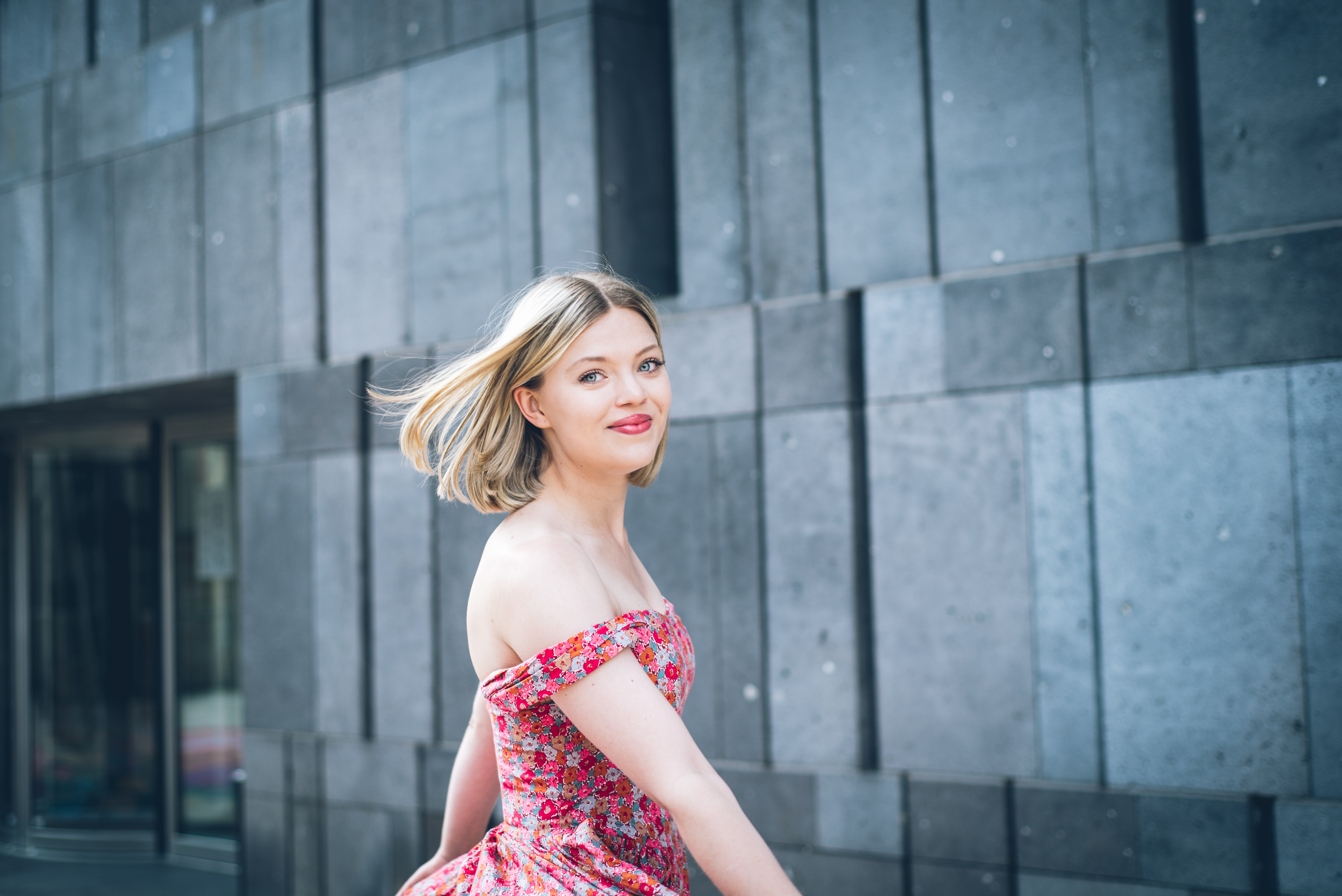 Eine lächelnde Frau in einem geblümten, schulterfreien Kleid steht im Freien vor einer modernen grauen Steinmauer; ihr blondes Haar weht sanft im Wind, während sie sich der Kamera zuwendet.
