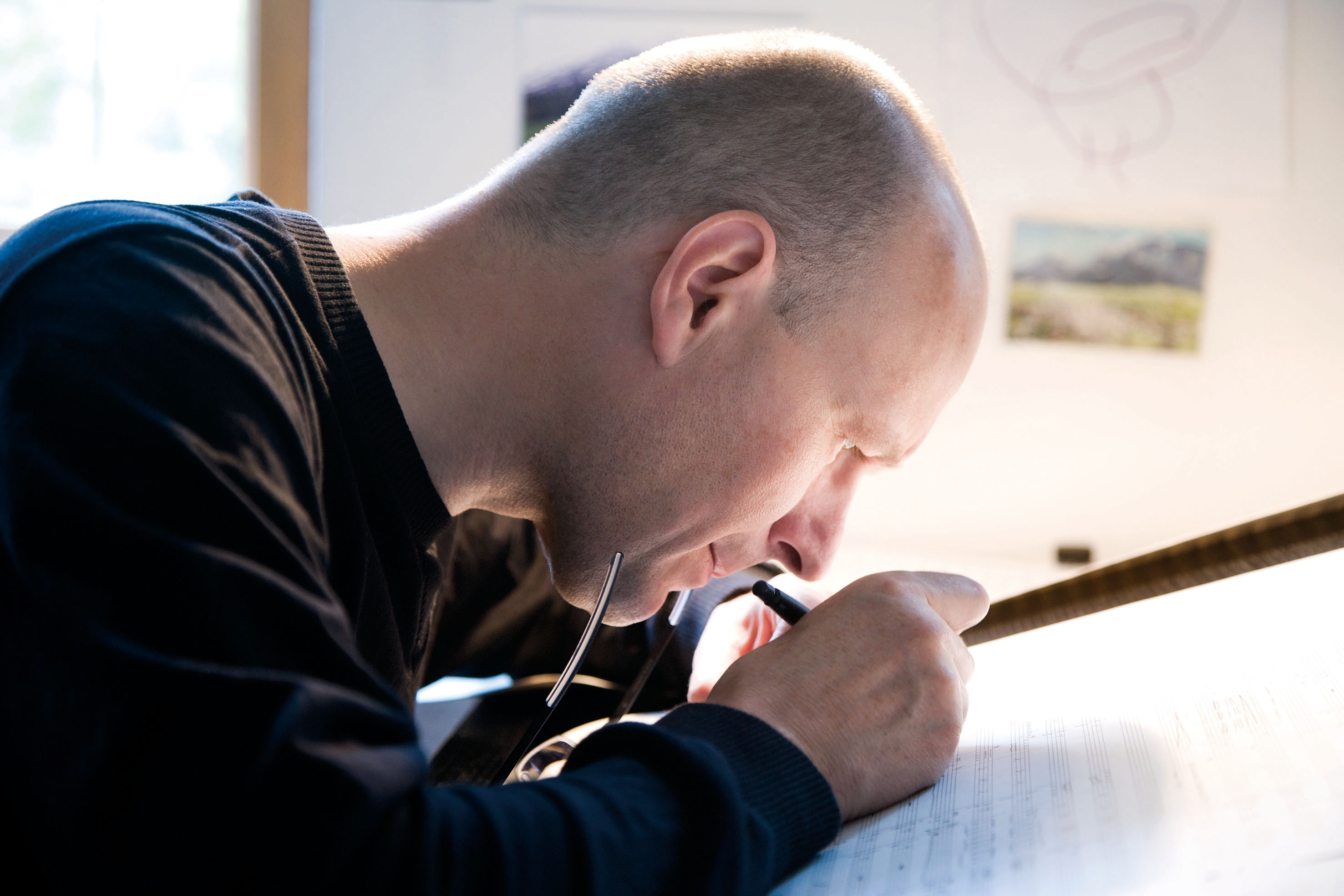 Eine Person mit Glatze sitzt an einem gut beleuchteten Schreibtisch und schreibt oder zeichnet konzentriert auf ein großes Blatt Papier, wobei sie in der einen Hand eine Brille und in der anderen einen Stift hält.