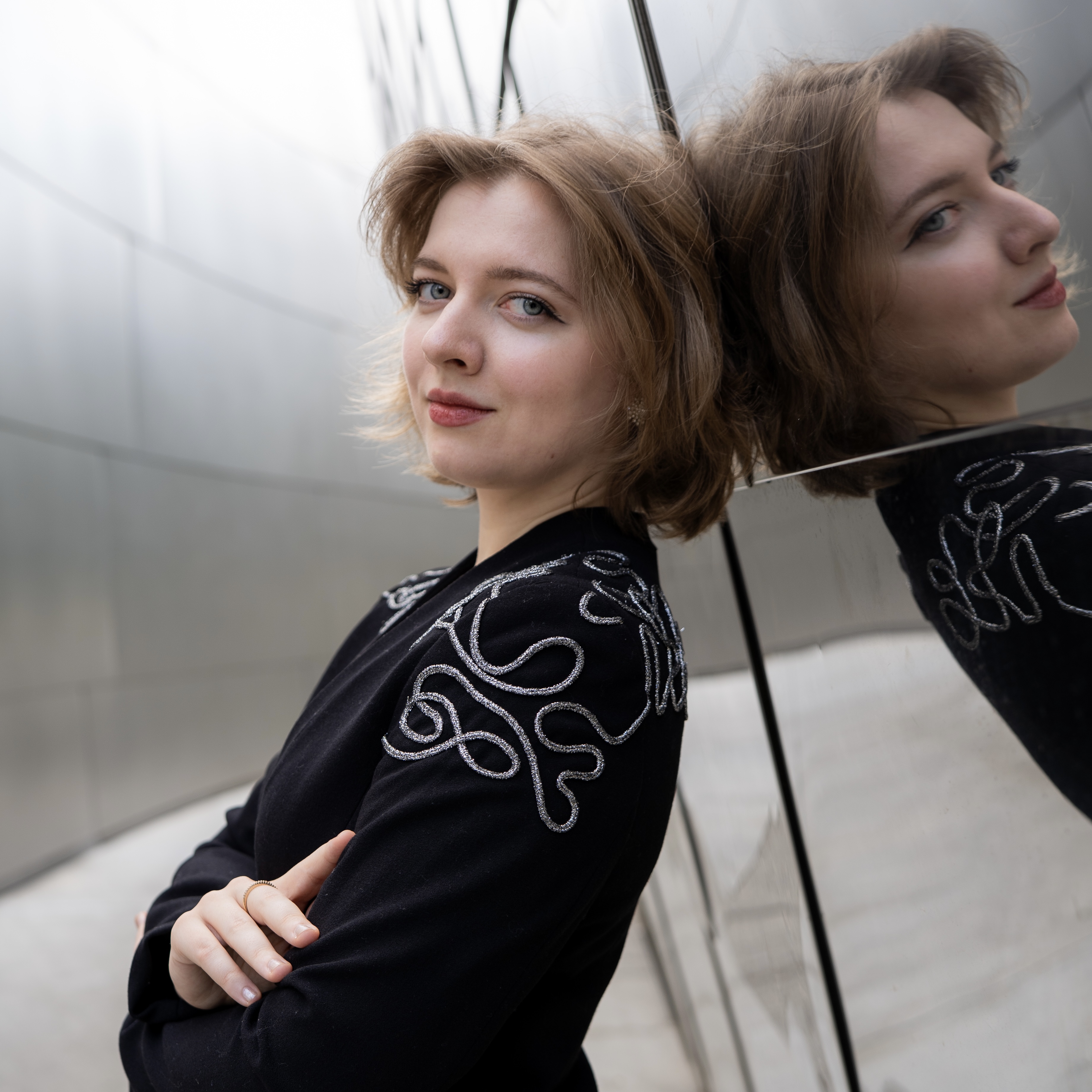 A person in a black jacket with decorative stitching leans against a shiny, curved metal wall, reflecting their face and upper body. They look at the camera with a calm expression.