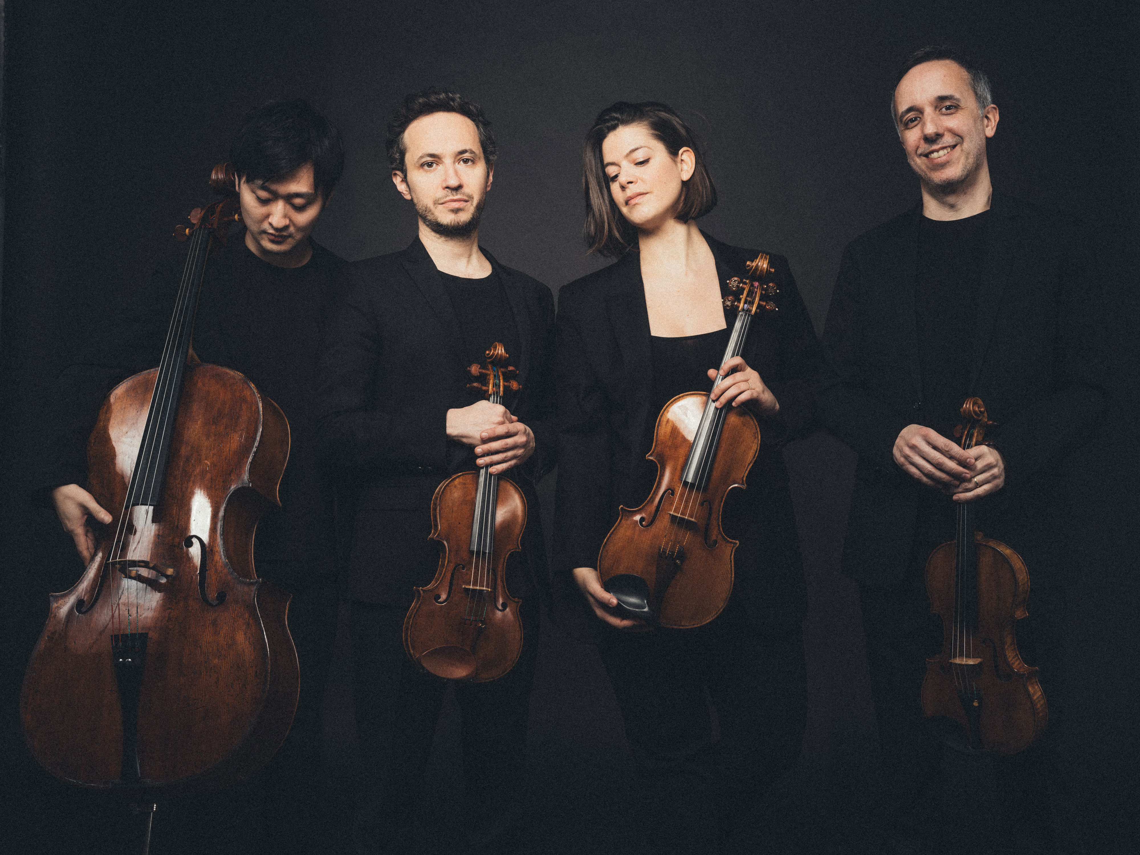 Vier Musiker posieren mit Streichinstrumenten vor dunklem Hintergrund. Sie tragen schwarze Abendgarderobe und halten ein Cello, zwei Geigen und eine Bratsche in den Händen. Sie stehen eng beieinander und blicken mit entspanntem Gesichtsausdruck in die Kamera.