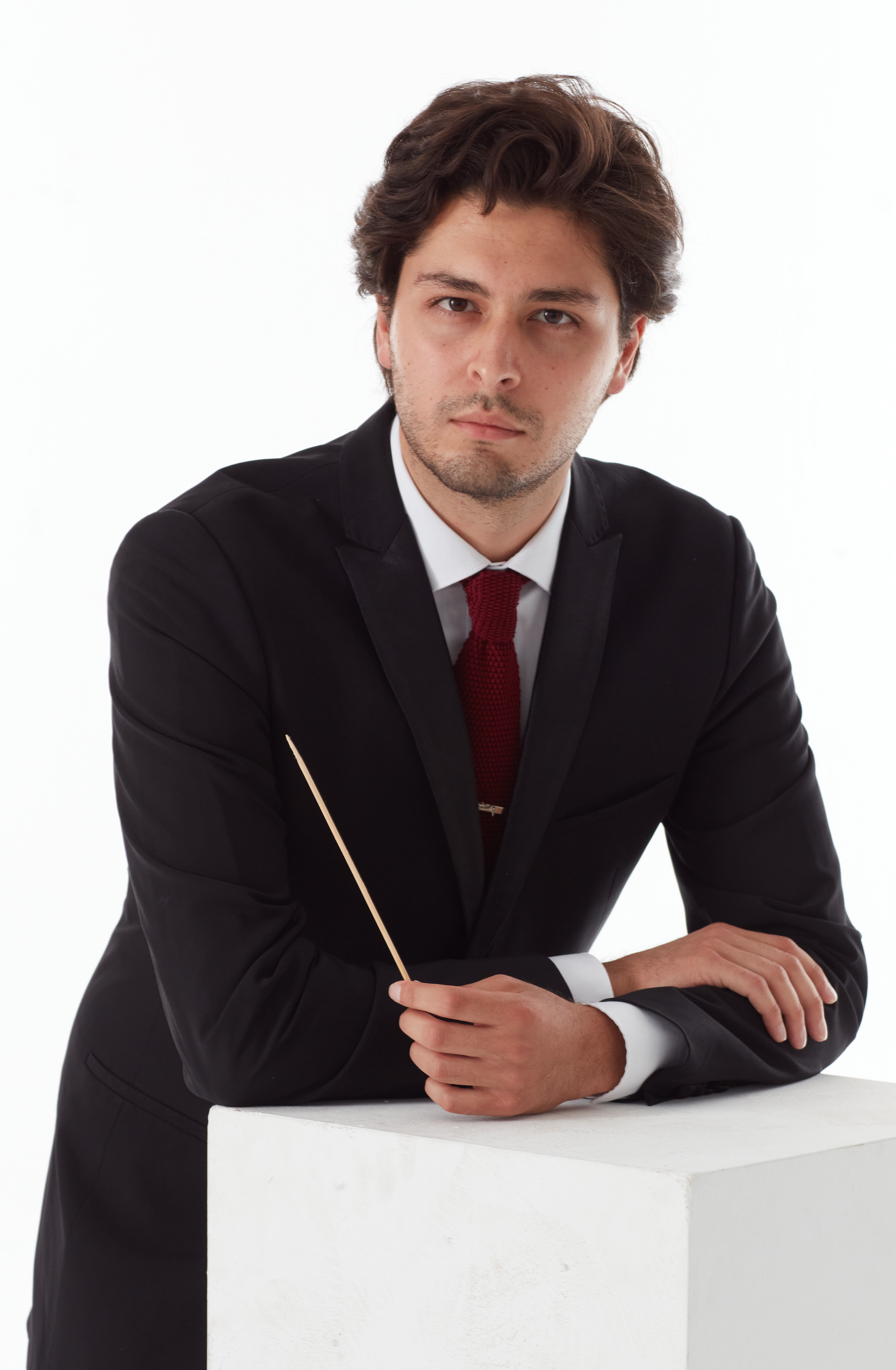 A man in a black suit and red tie leans on a white block, holding a thin baton. He has medium-length brown hair and a serious expression against a plain white background.