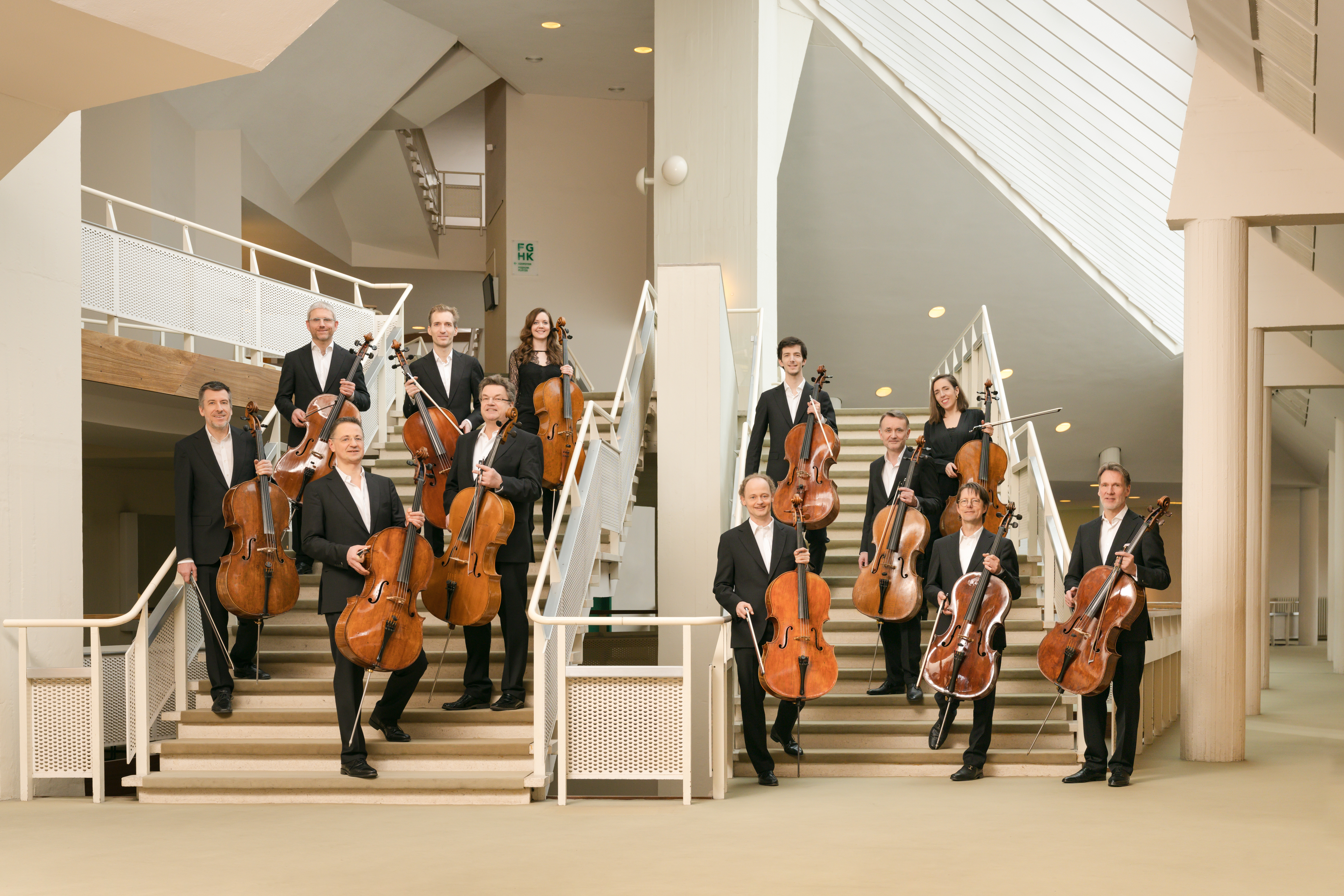 Die zwölf Cellisten stehen in schwarzer Konzertkleidung auf der Treppe in der Philharmonie mit ihren Instrumenten in der Hand.