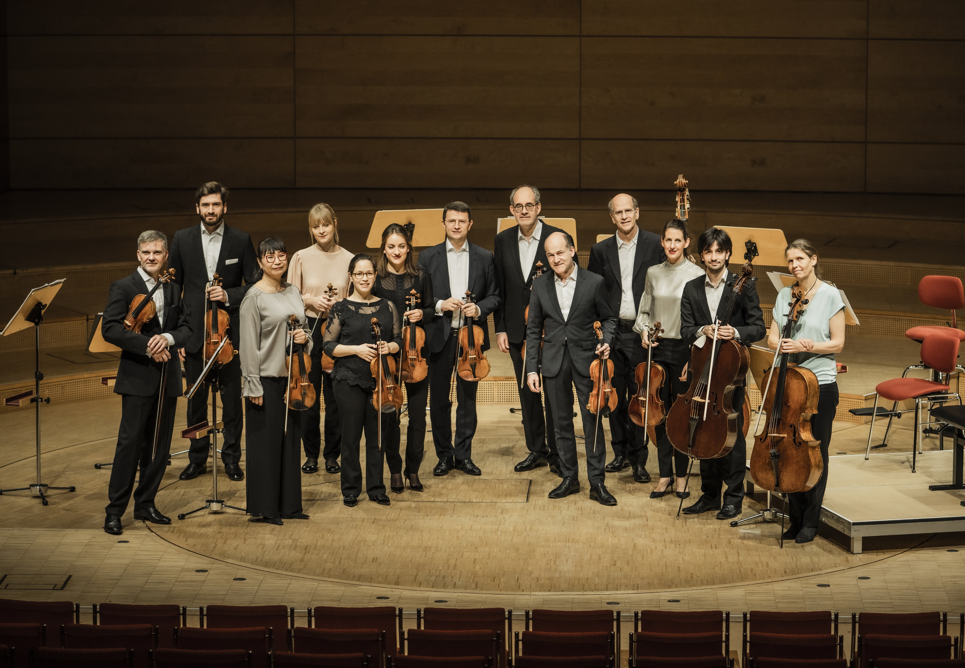 Zwölf Musiker in förmlicher Kleidung stehen auf einer hölzernen Bühne in einem Konzertsaal und halten Streichinstrumente, darunter Geigen, Bratschen und Celli, vor leeren Notenständern und Stühlen im Hintergrund.