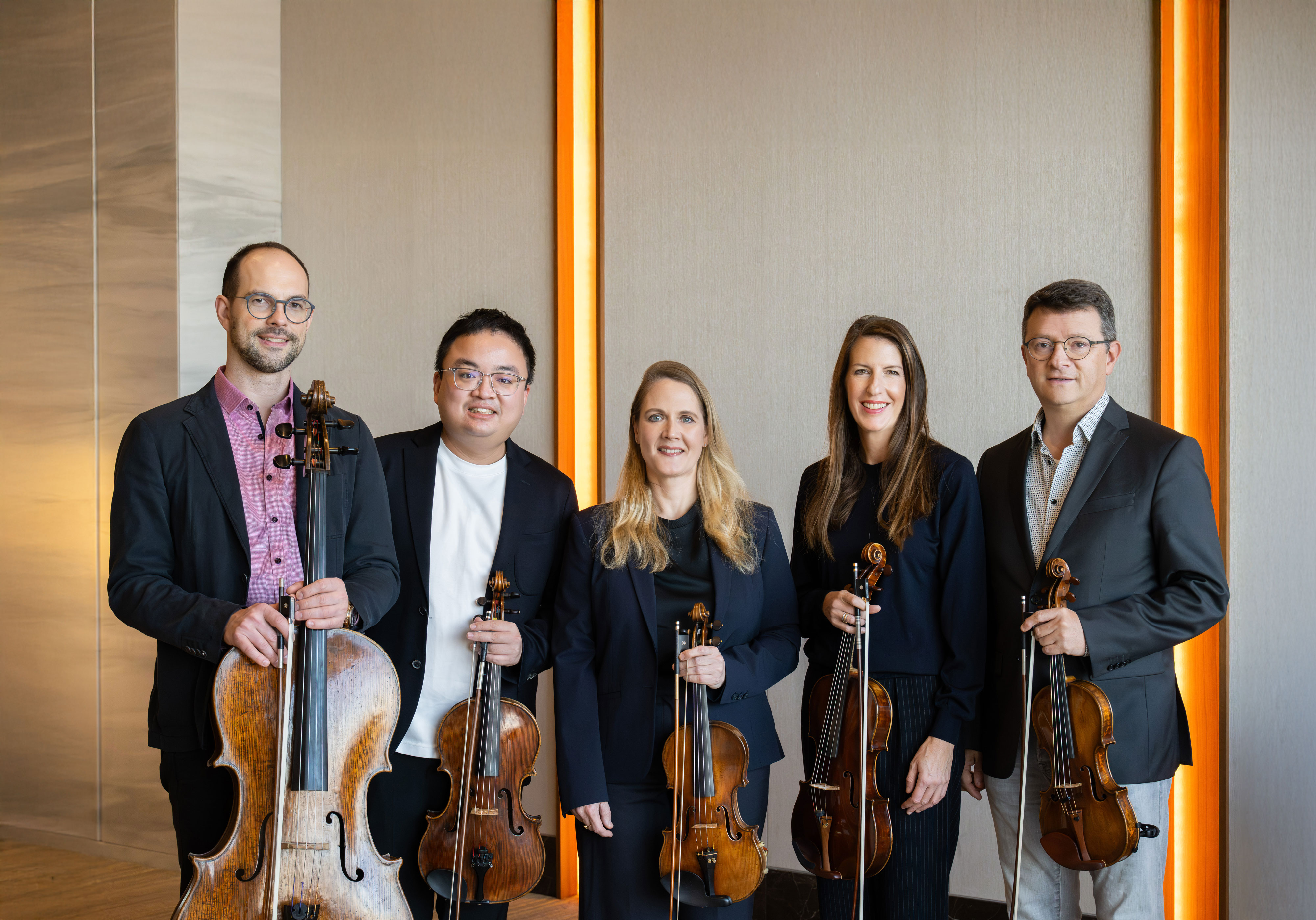 Fünf Musiker stehen in einem Innenraum nebeneinander, halten Streichinstrumente in der Hand, darunter Geigen, eine Bratsche und ein Cello, alle in Geschäfts- oder Konzertkleidung, und lächeln in die Kamera.