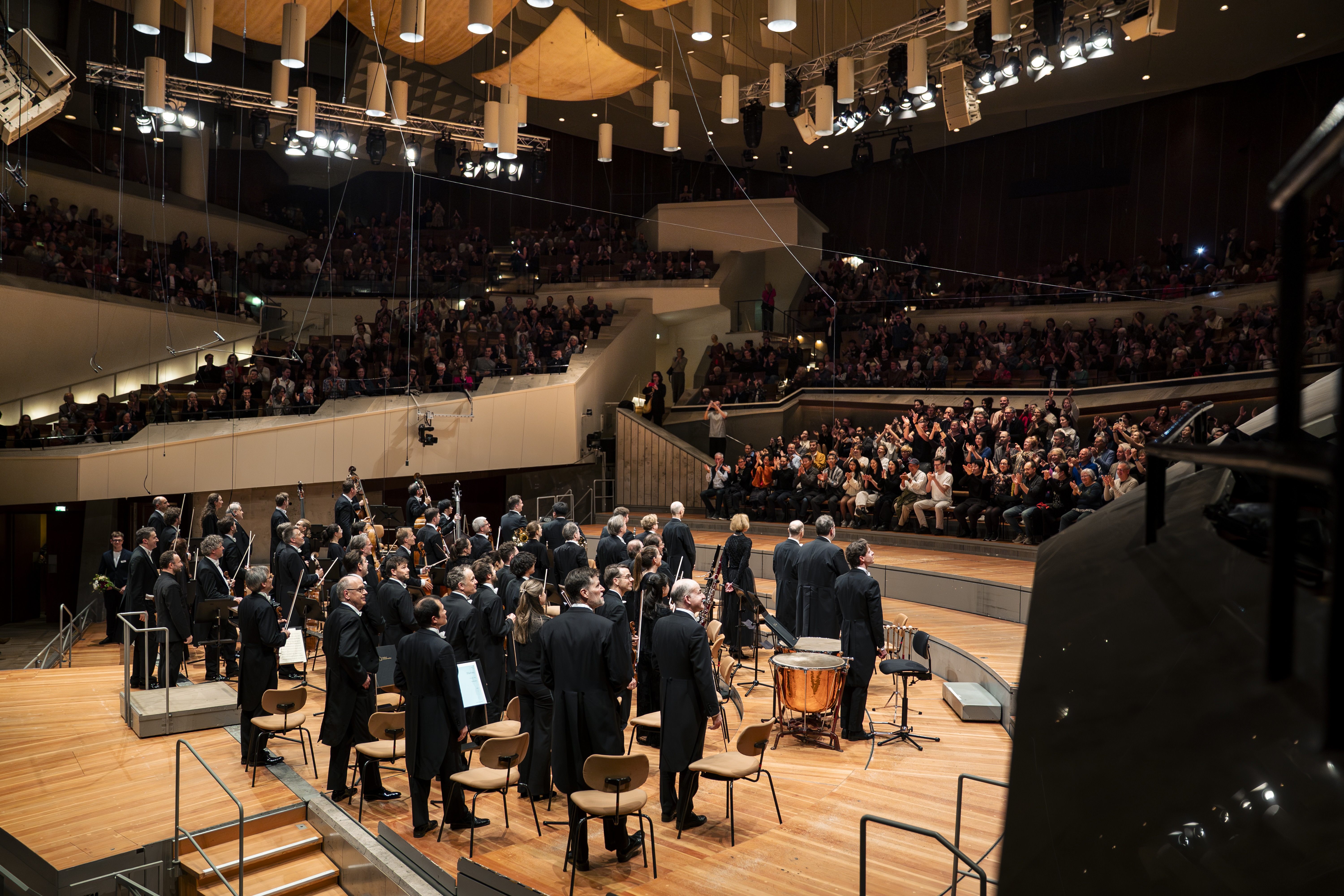 Ein Orchester steht auf der Bühne vor einem großen Publikum in einem modernen Konzertsaal. Das Publikum sitzt in Reihen und spendet den Musikern herzlichen Applaus. Der Saal ist durch Deckenbeleuchtung hell ausgeleuchtet.