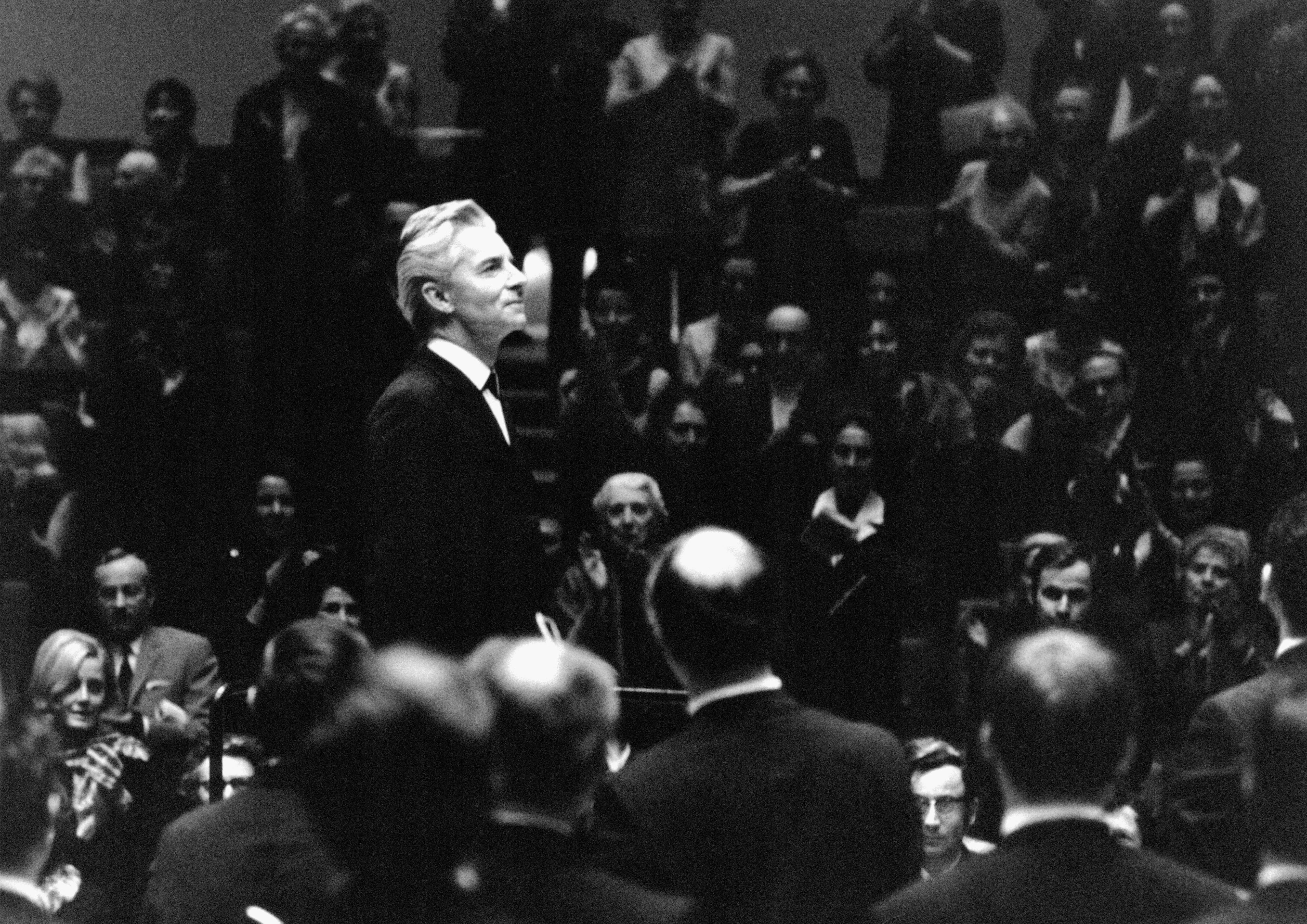 Black and white photograph of Herbert von Karajan ca. 1970, standing in front of orchestra and applauding audience.