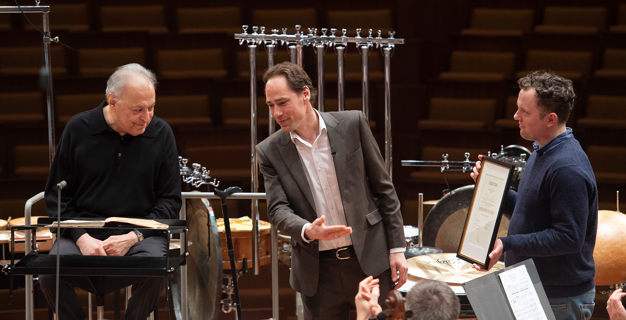 Drei Männer stehen in einem Konzertsaal auf der Bühne; einer sitzt an einem Notenpult, einer im Anzug gestikuliert zu ihm, und einer im Pullover hält eine gerahmte Urkunde. Im Hintergrund sind Perkussionsinstrumente zu sehen.