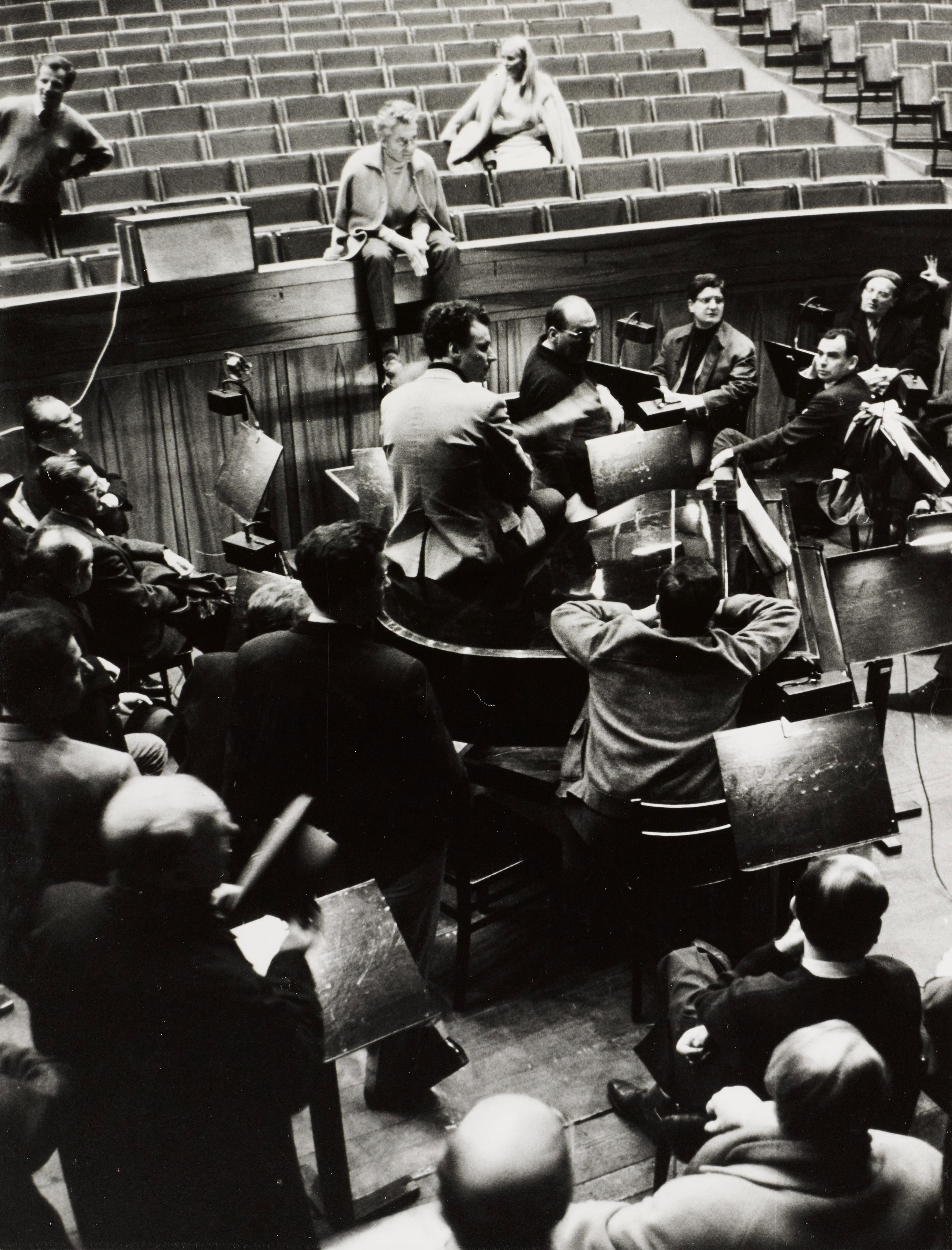 A black and white photo of an orchestra rehearsal. Musicians sit with their instruments. Conductor Herbert von Karajan sits casually on the balustrade. The hall is largely empty, apart from a few people sitting scattered around.