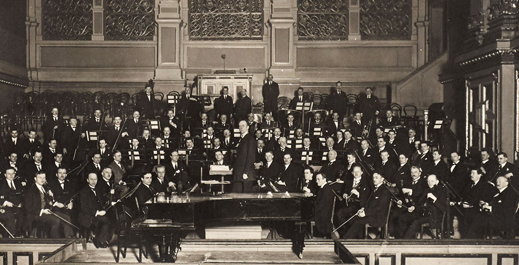 Auf der Bühne sitzt ein großes Orchester von Männern in formeller Kleidung mit ihren Musikinstrumenten, die um einen Flügel herum angeordnet sind. Der Dirigent Wilhelm Furtwängler steht in der Mitte. Im Hintergrund sind verzierte Wände und leere Stühle zu sehen.