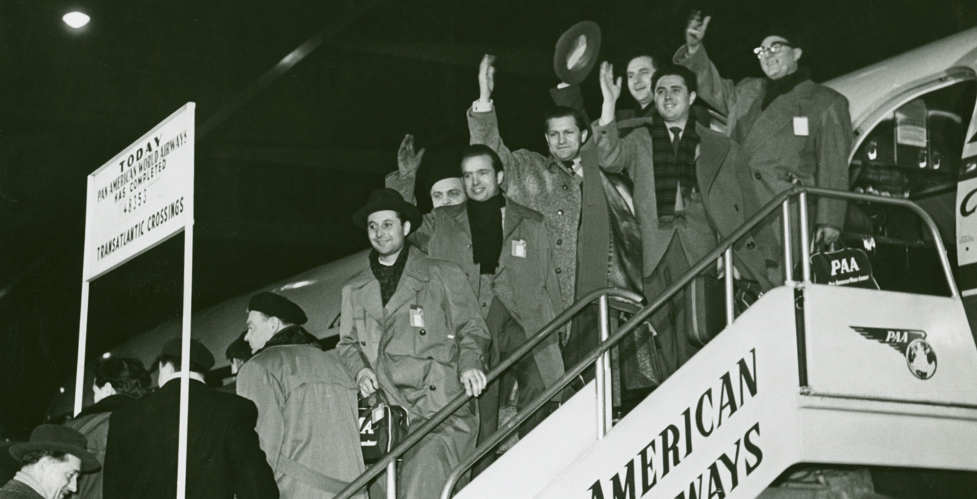 Black and white photo: Orchestra members standing on the gangway of an American Airways aeroplane and waving.