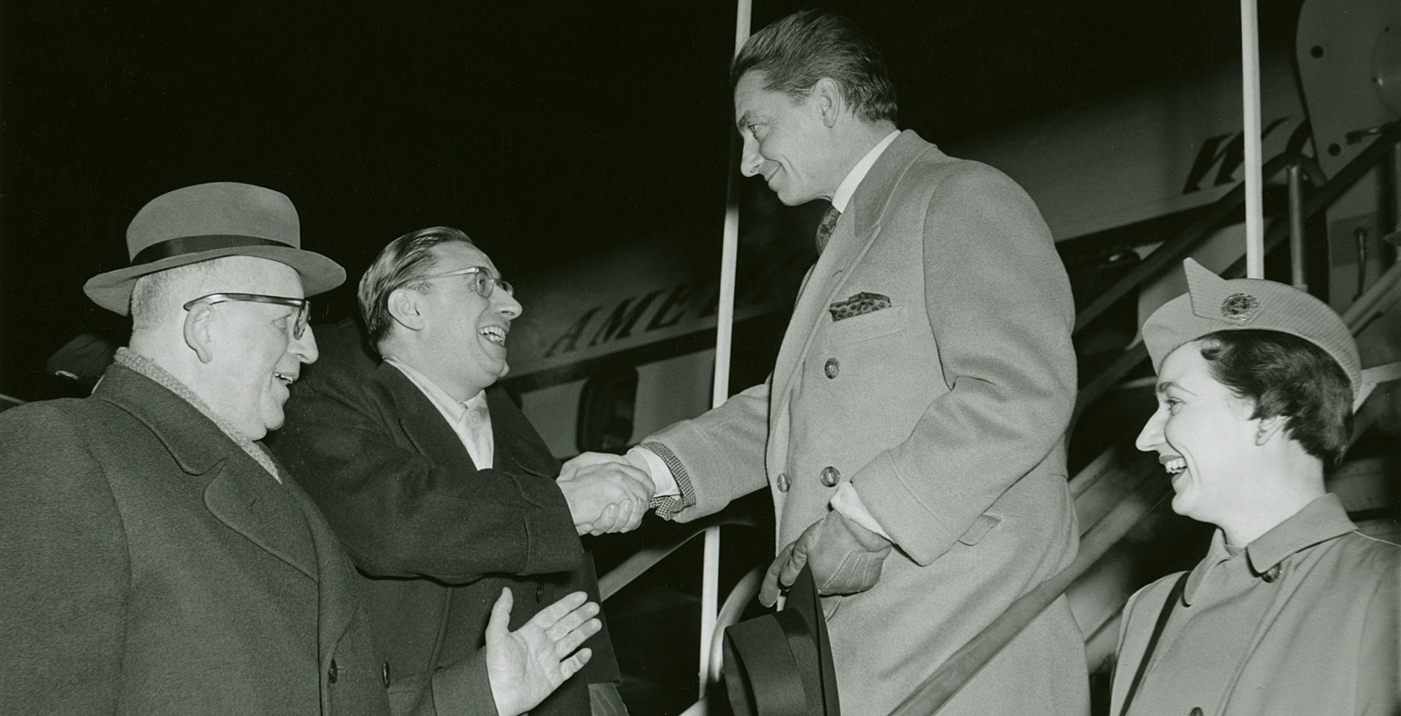 Herbert von Karajan shakes hands with a man, a second man stands next to him. A flight attendant can be seen on the right.