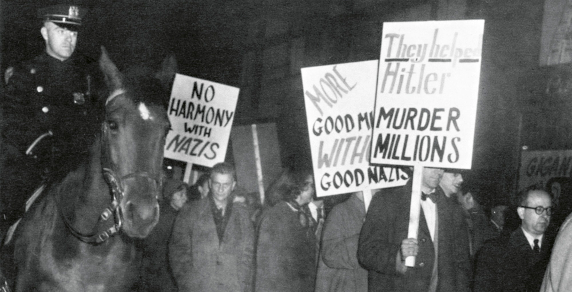 A police officer on horseback looks at protesters holding up signs. Among other things, they read “No harmony with Nazis”.
