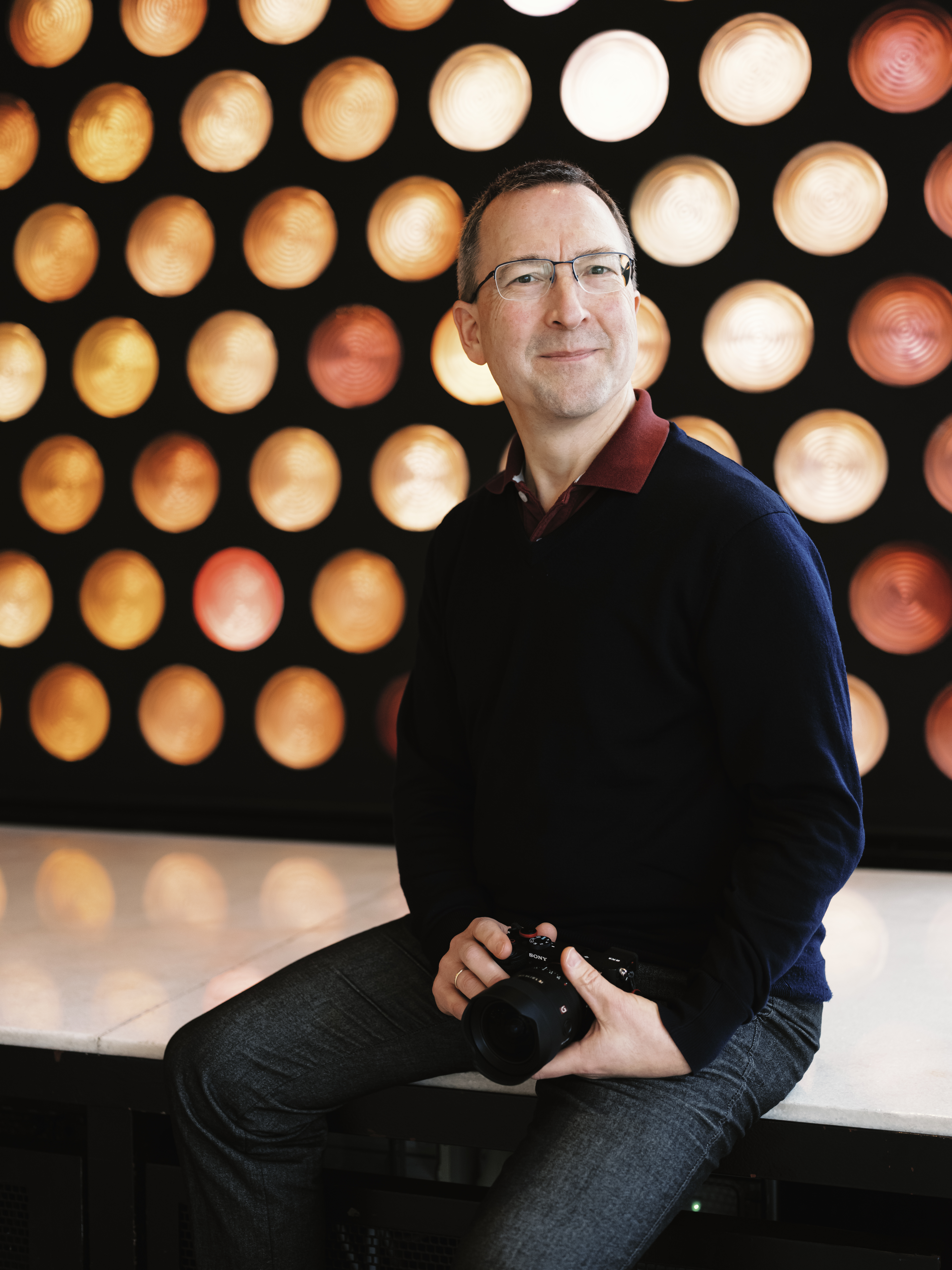 Ein Mann mit Brille und einem dunklen Pullover sitzt mit einer Kamera in der Hand vor einer Wand mit runden, orange- und gelb leuchtenden Lichtern.