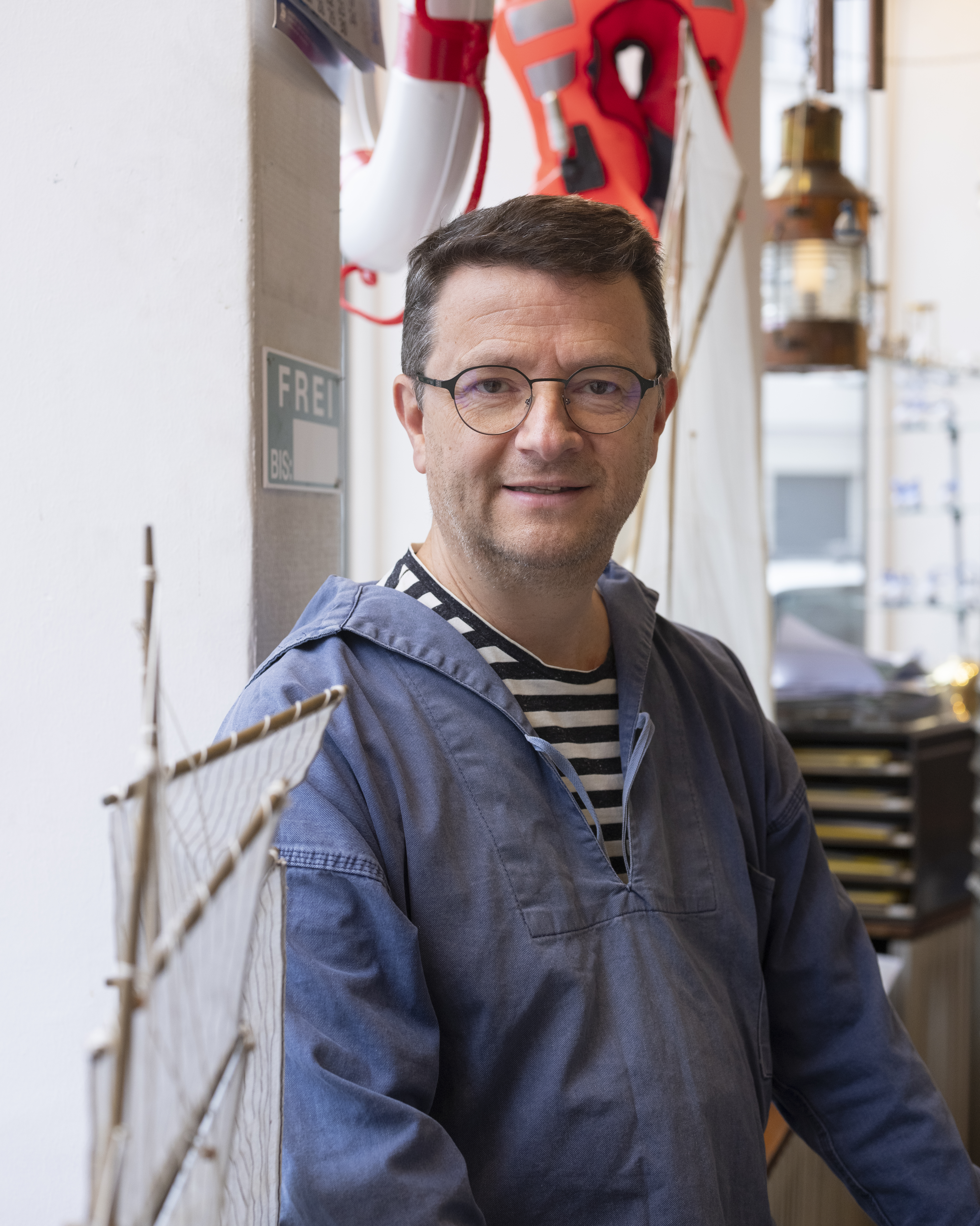 A man wearing glasses, a blue jacket, and a striped shirt stands indoors next to a model ship. Nautical decor, including a life preserver and lantern, is visible in the background.