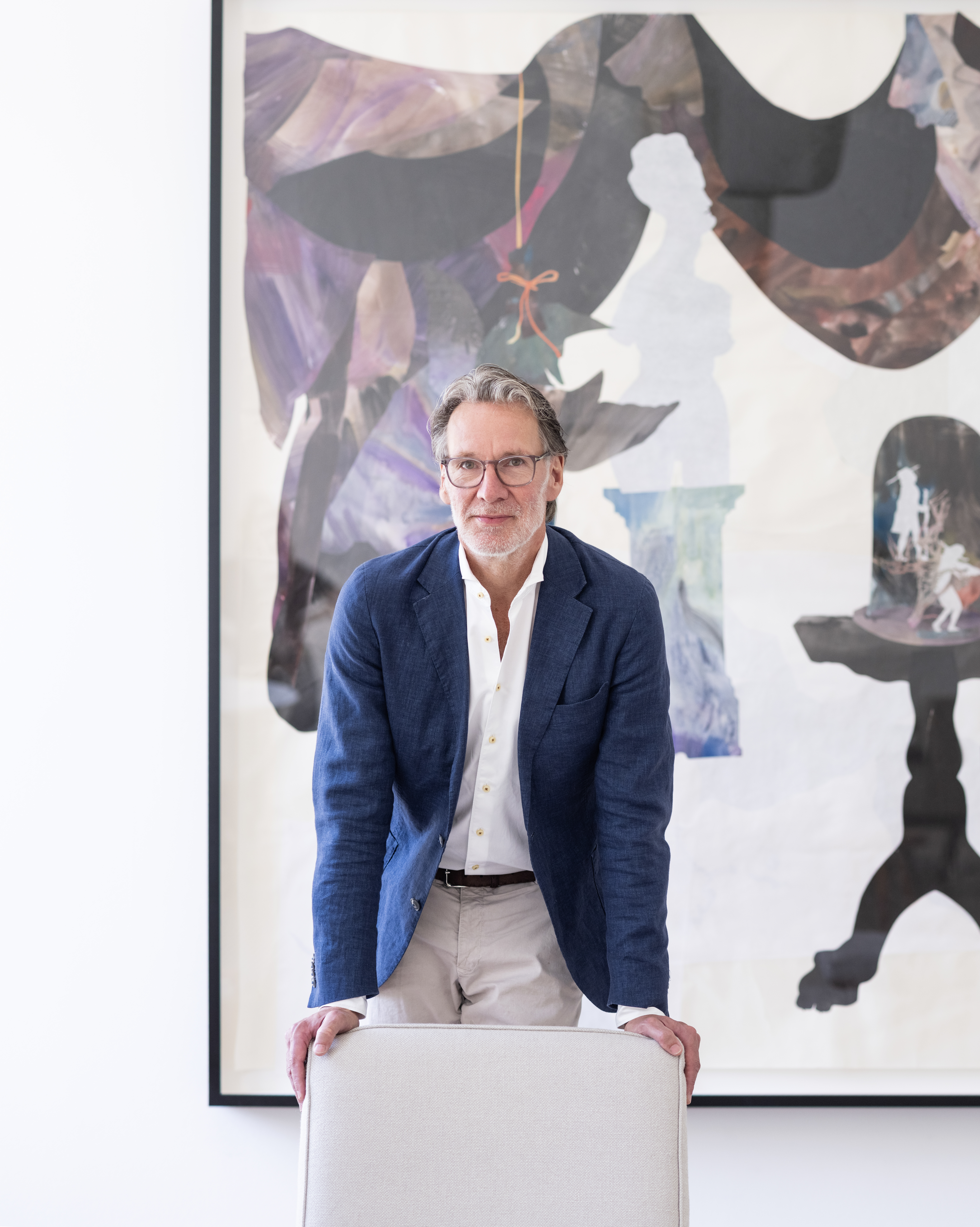 A man in a blue blazer and glasses stands behind a light chair, with a large abstract artwork featuring silhouettes and dark drapery in the background.