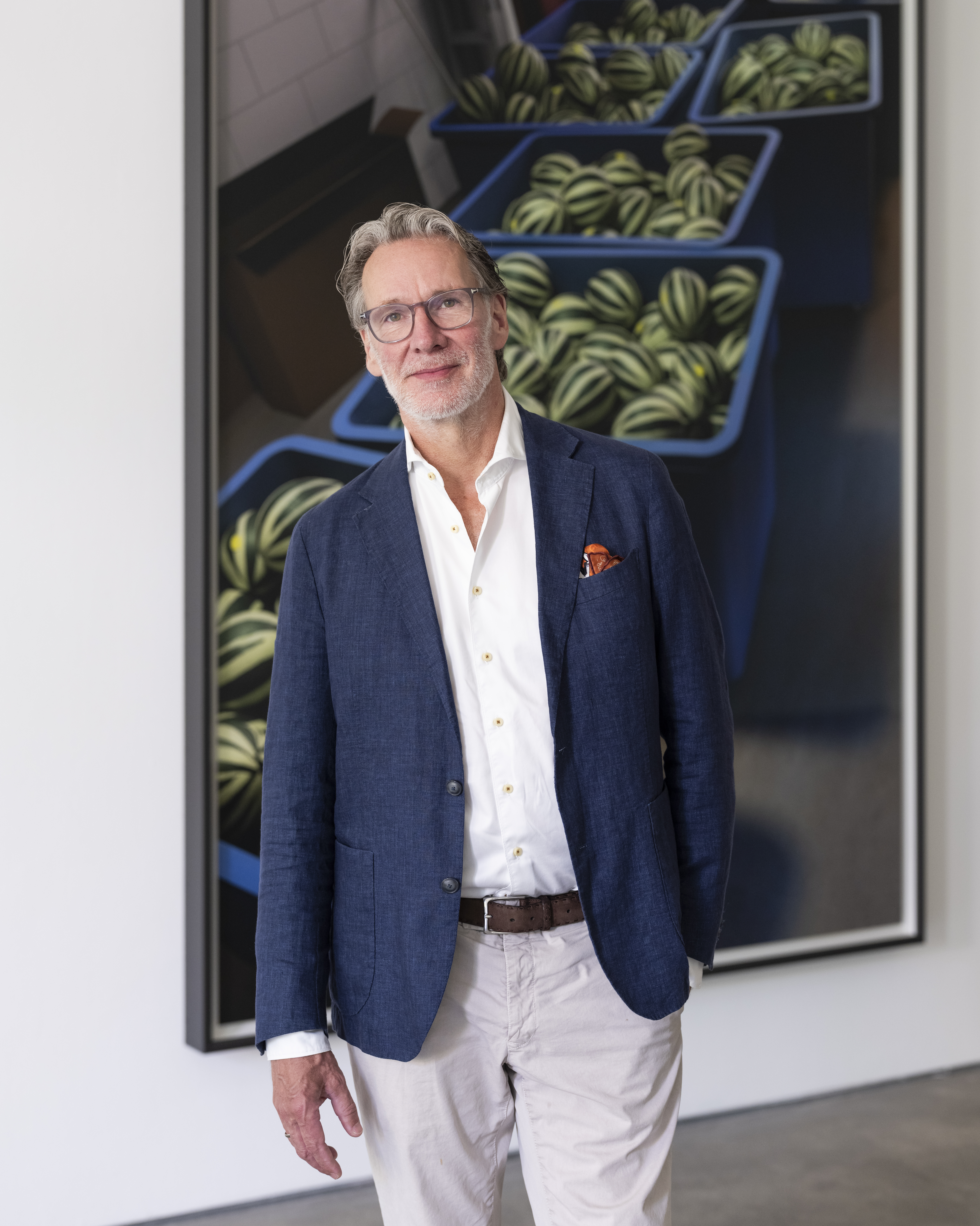 Ein Mann mit Brille, blauem Blazer und khakifarbener Hose steht im Raum und lächelt. Hinter ihm ist ein großes Kunstwerk zu sehen, das mehrere blaue Behälter zeigt, die mit kleinen gestreiften Wassermelonen gefüllt sind.