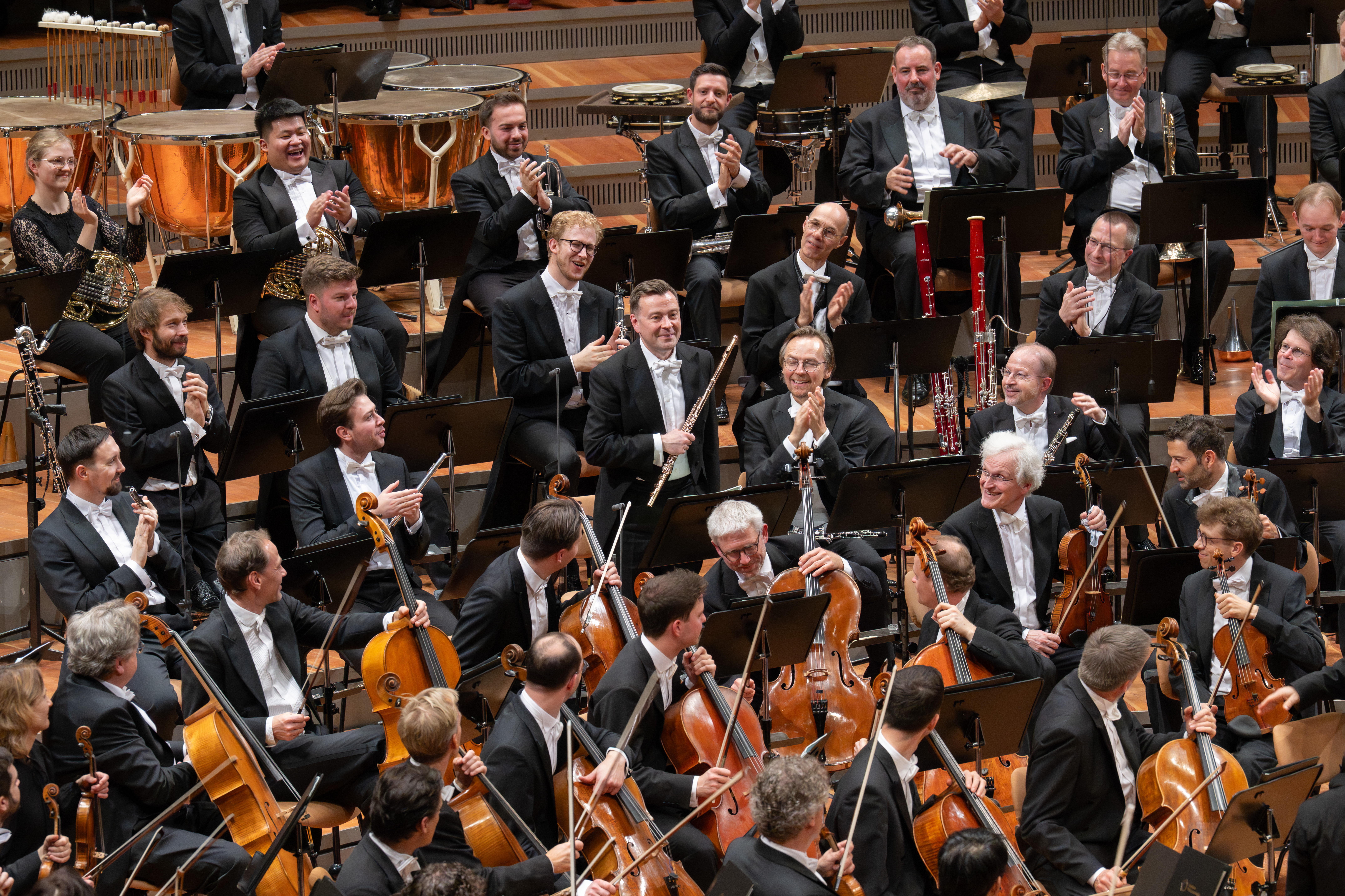 A large orchestra of musicians in formal black attire sit onstage with string, brass, and wind instruments, smiling and applauding in a concert hall.