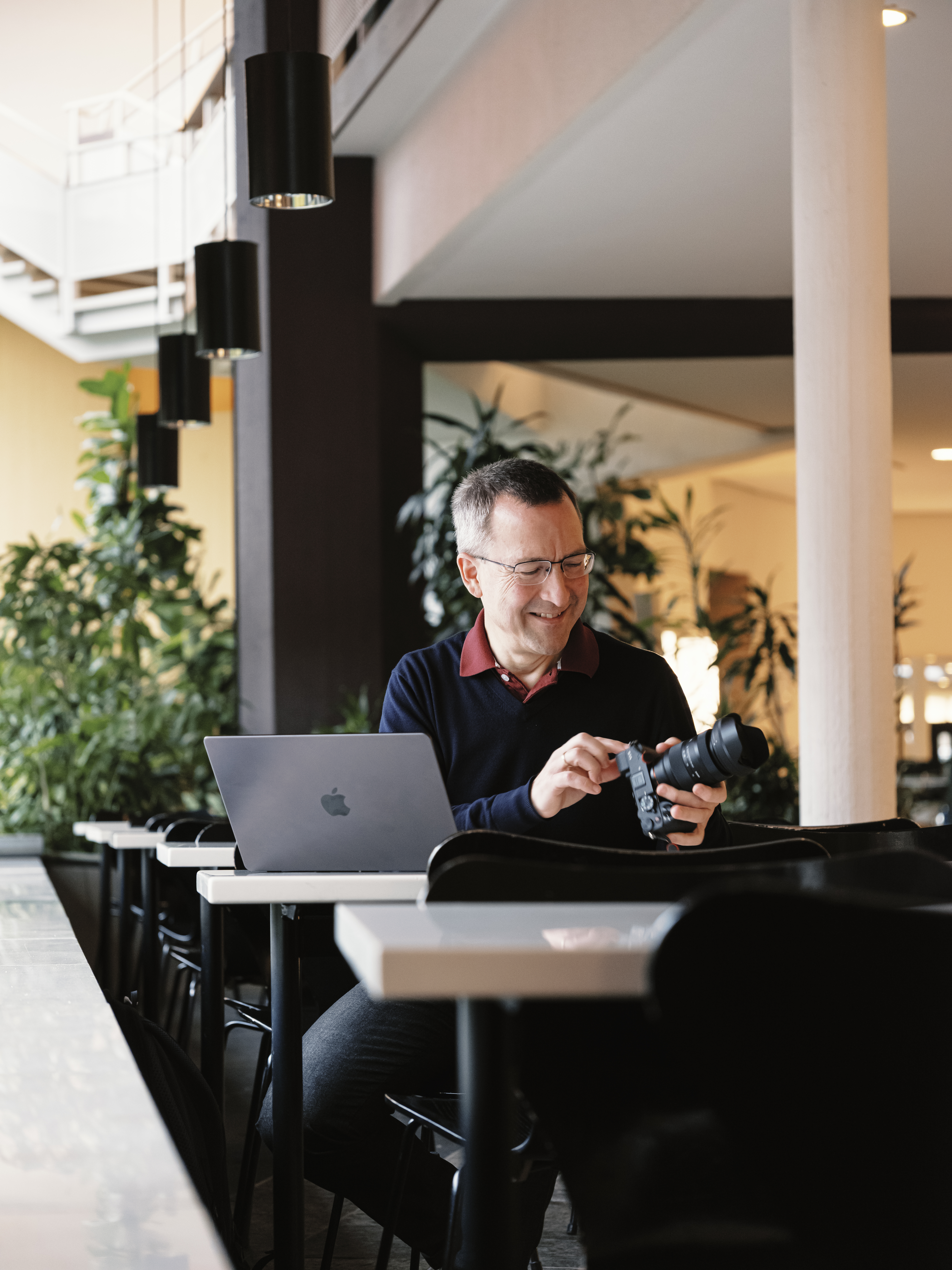 Ein Mann sitzt mit einem Laptop an einem Tisch und lächelt, während er eine Kamera einstellt. Die Kulisse scheint ein moderner Innenraum zu sein, im Hintergrund sind Pflanzen und Hängelampen zu sehen.