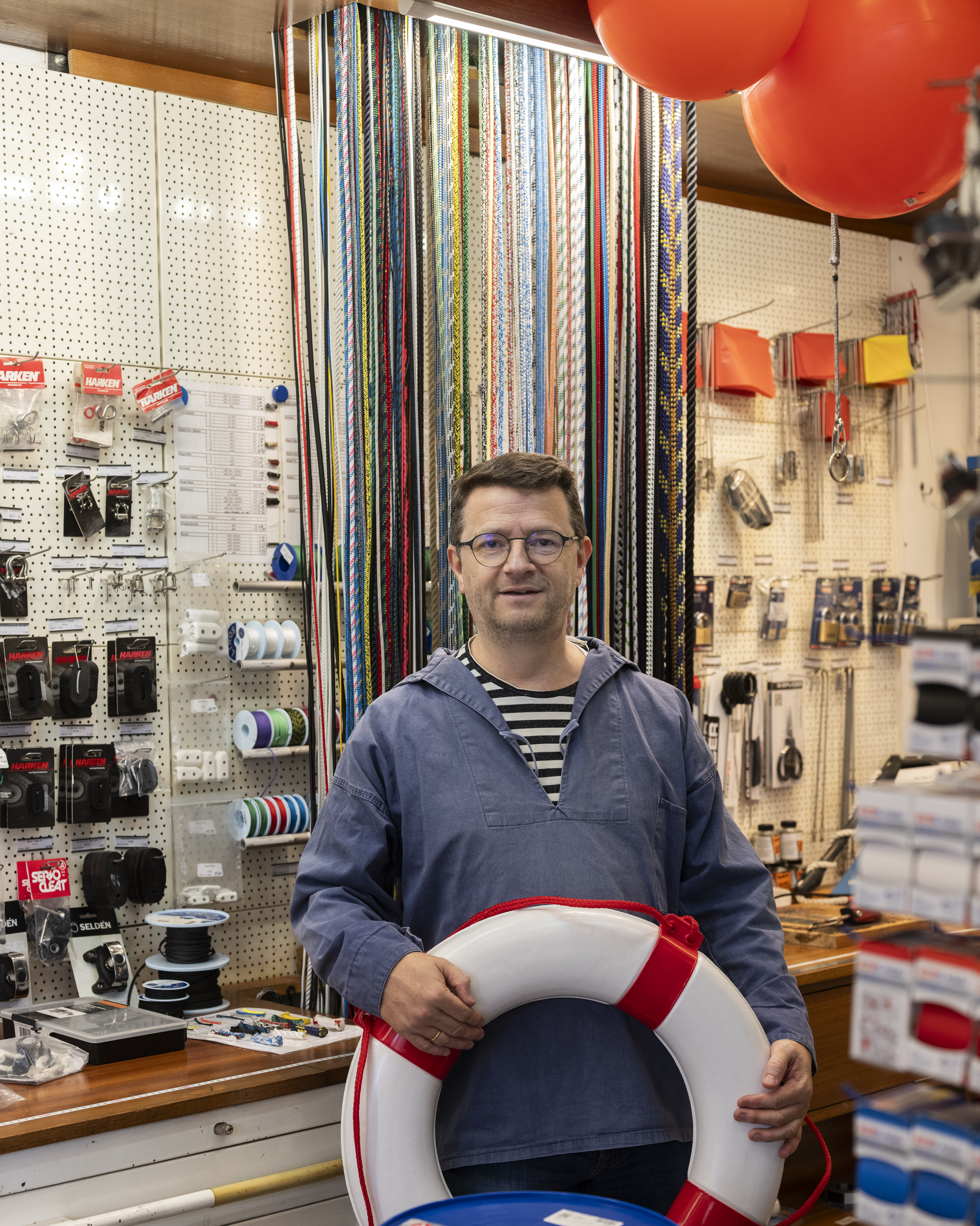Ein Mann mit Brille und blauem Hemd hält eine Rettungsweste in der Hand. Hinter ihm sind bunte Seile, Werkzeuge und Baumaterialien zu sehen, und über ihm hängen rote Luftballons.