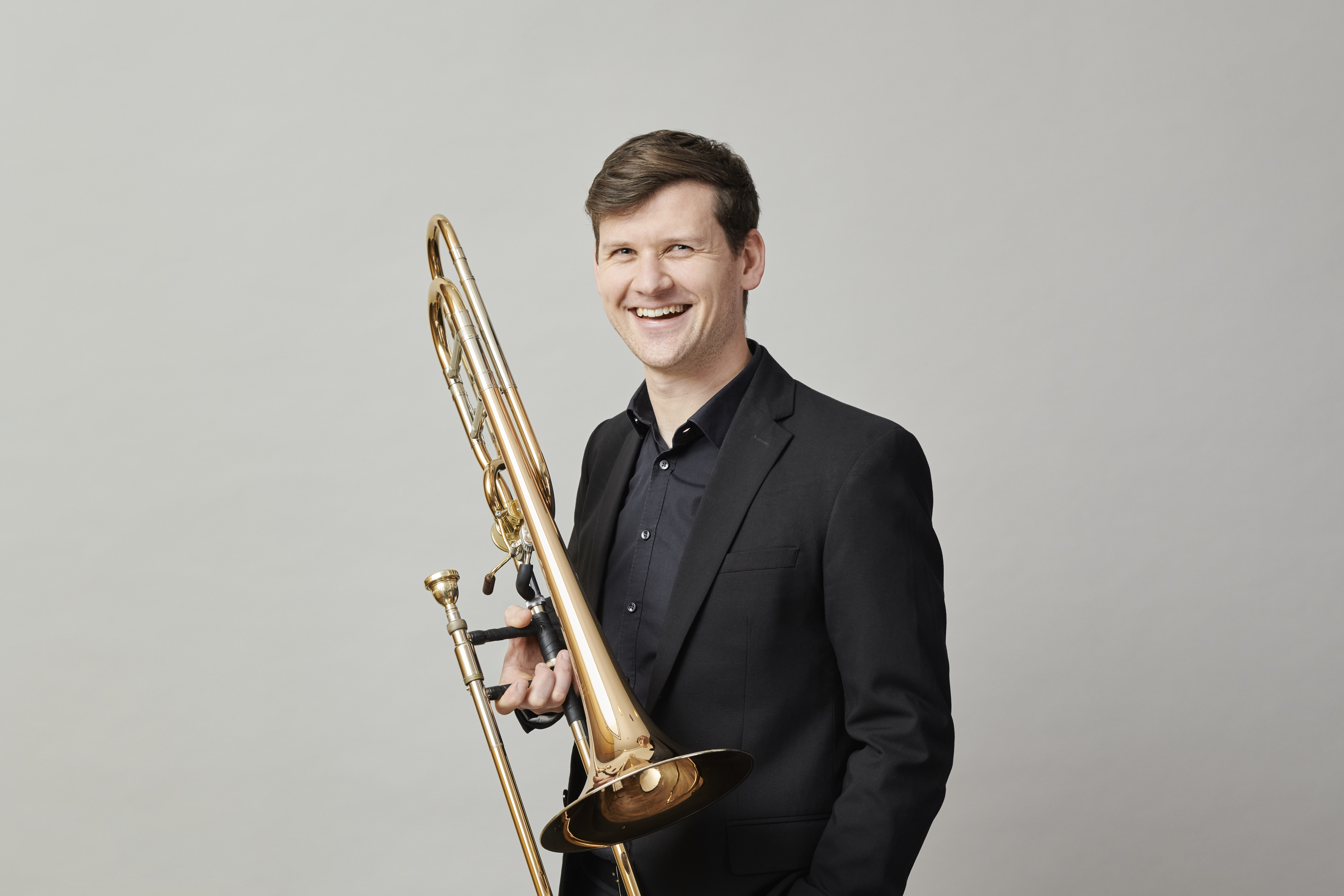 A smiling man in a black suit holds a trombone against a plain, light grey background.