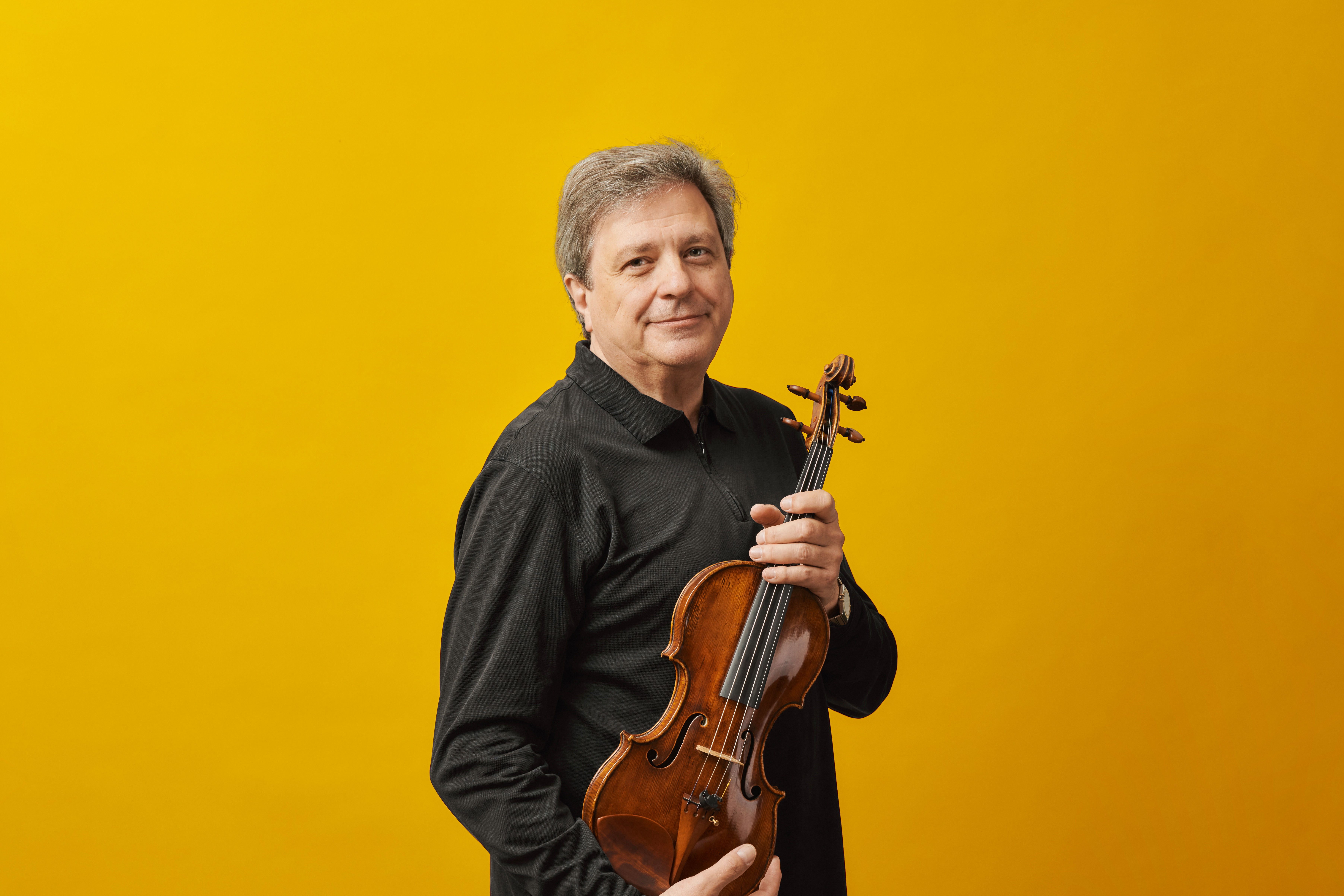 A middle-aged man with gray hair stands against a bright yellow background holding a violin.
