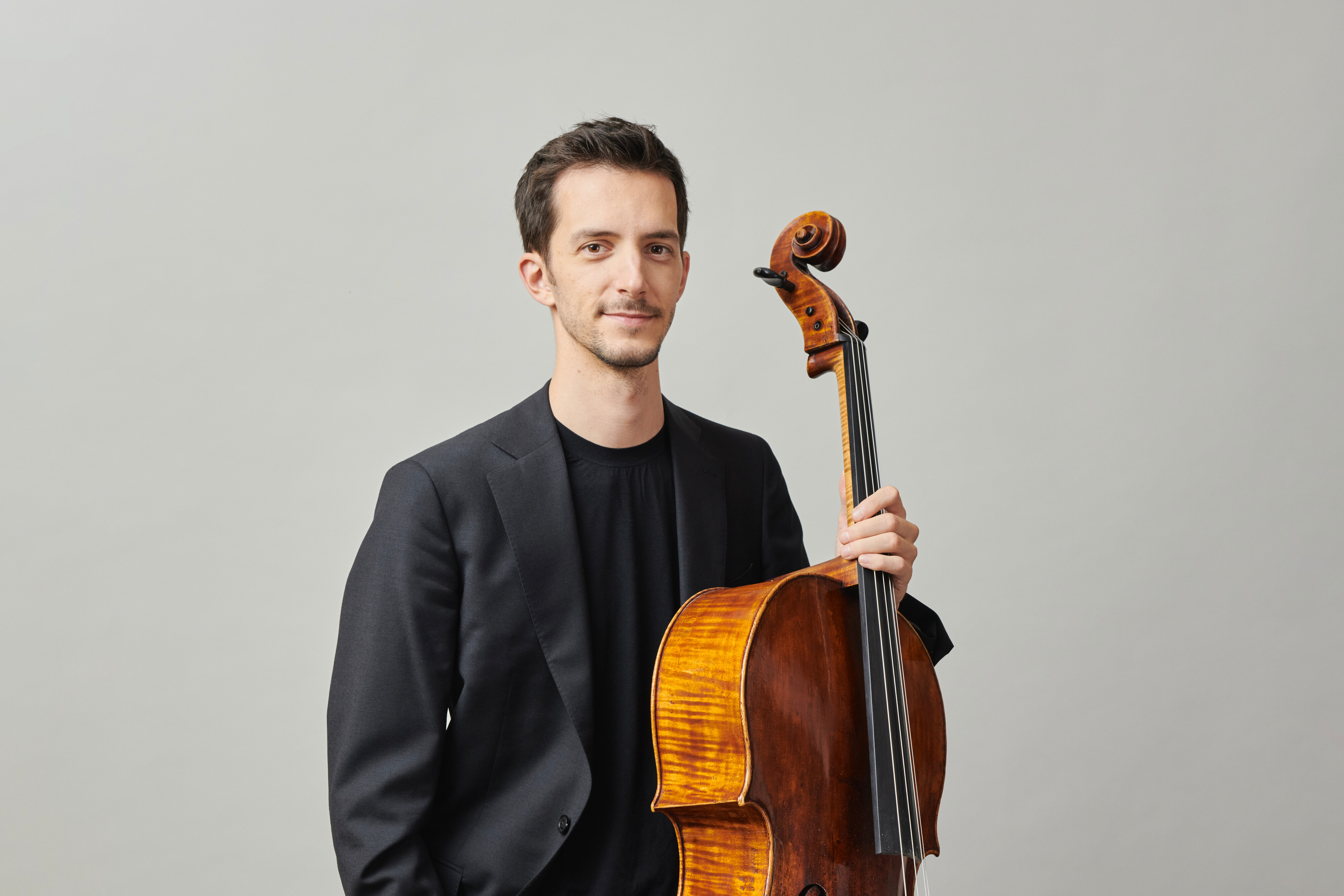 Bruno Delepelaire in a black suit stands holding a cello upright beside him against a plain light gray background, looking at the camera with a slight smile.