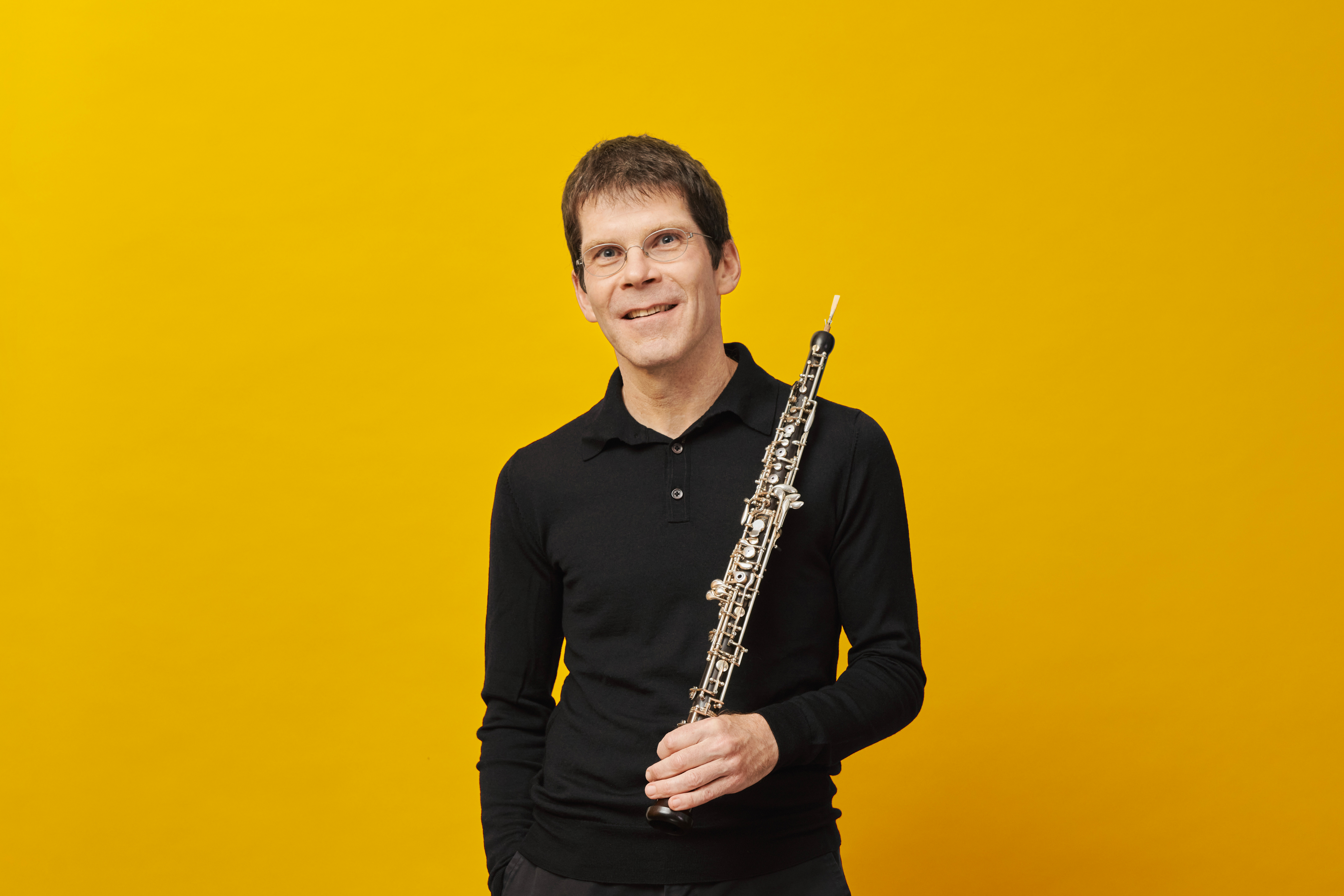 Ein Mann in einem schwarzen Hemd lächelt, während er eine Oboe vor einem einfarbig gelben Hintergrund hält.