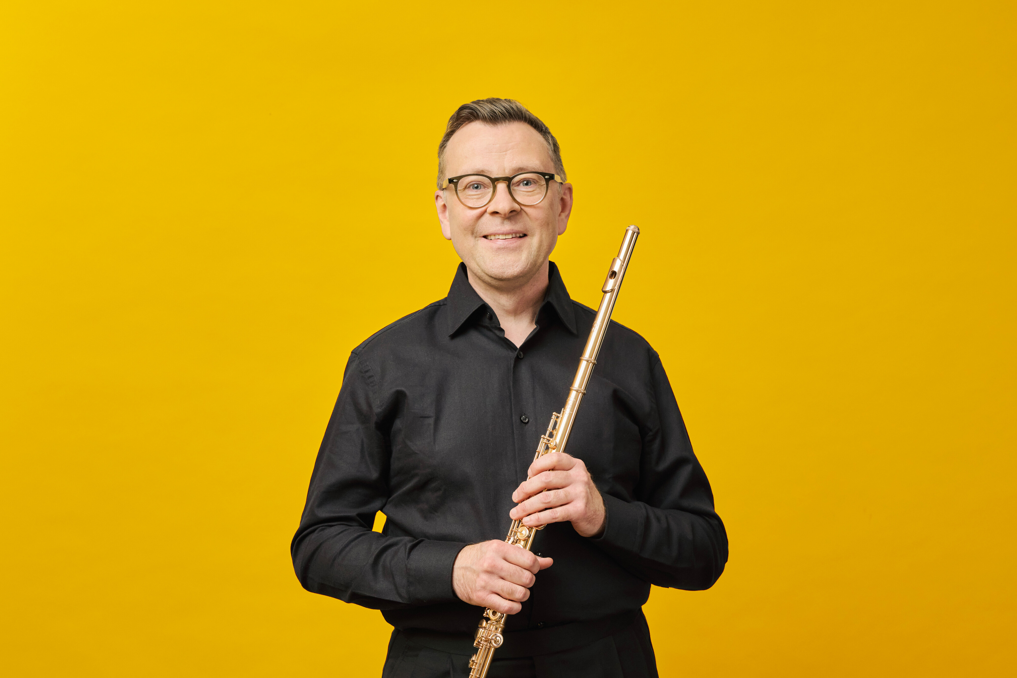 A smiling Stefán Ragnar Höskuldsson in glasses and a black shirt holds a flute upright in front of a bright yellow background.